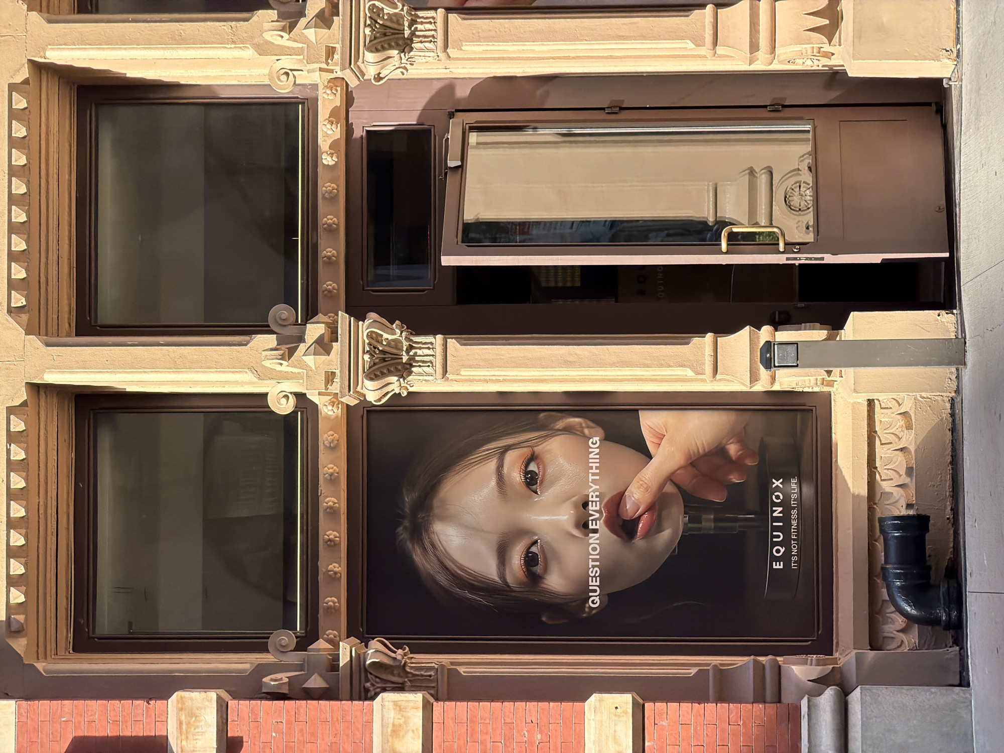 The image depicts the facade of a building, likely in New York City, United States, based on architectural style. The building features tan and brown stone elements with intricate detailing. There are two windows visible, both framed with ornate trim. Below the lower window is a wall made of red bricks.
An advertisement for Equinox, a fitness company, is positioned between the windows. The ad features a person's face and a hand holding a dumbbell up to their mouth with the tagline "Question Everything. Equinox. It's not fitness. It's life." A black pipe is visible adjacent to the ad, mounted on the building's surface.