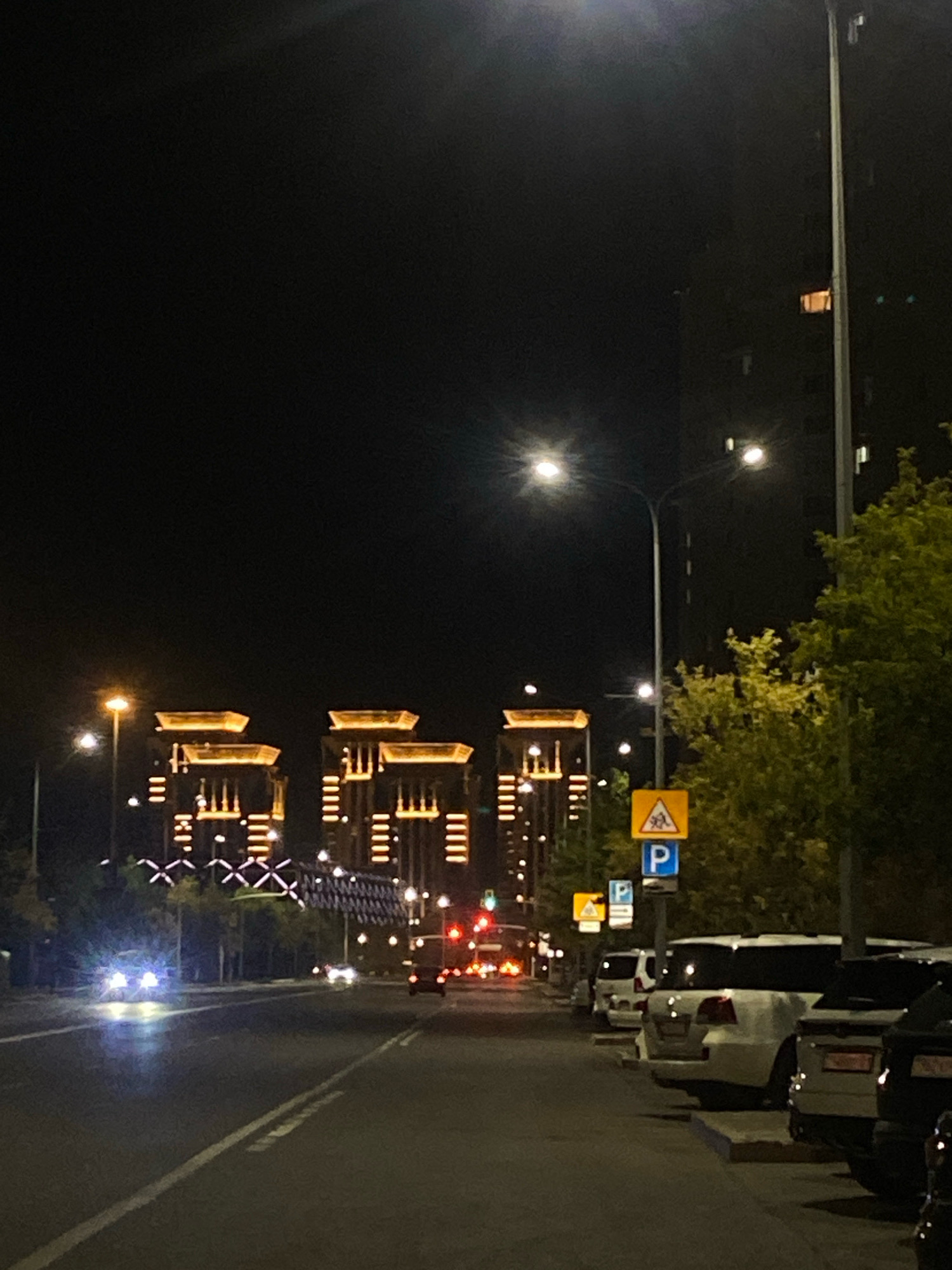 The image shows a street scene at night in Astana, Kazakhstan. In the background, there are several illuminated buildings with a distinctive architectural style featuring golden tops. To the right, a tall lamppost stands next to a large tree with green foliage. Several cars are parked along the side of the road. A yellow traffic sign with a pedestrian symbol and a blue parking sign are visible on a pole. The street is paved and has white lane markings. Cars are driving on the road, with their headlights illuminating the scene. The overall impression is a modern urban environment at night, likely a commercial or residential area.