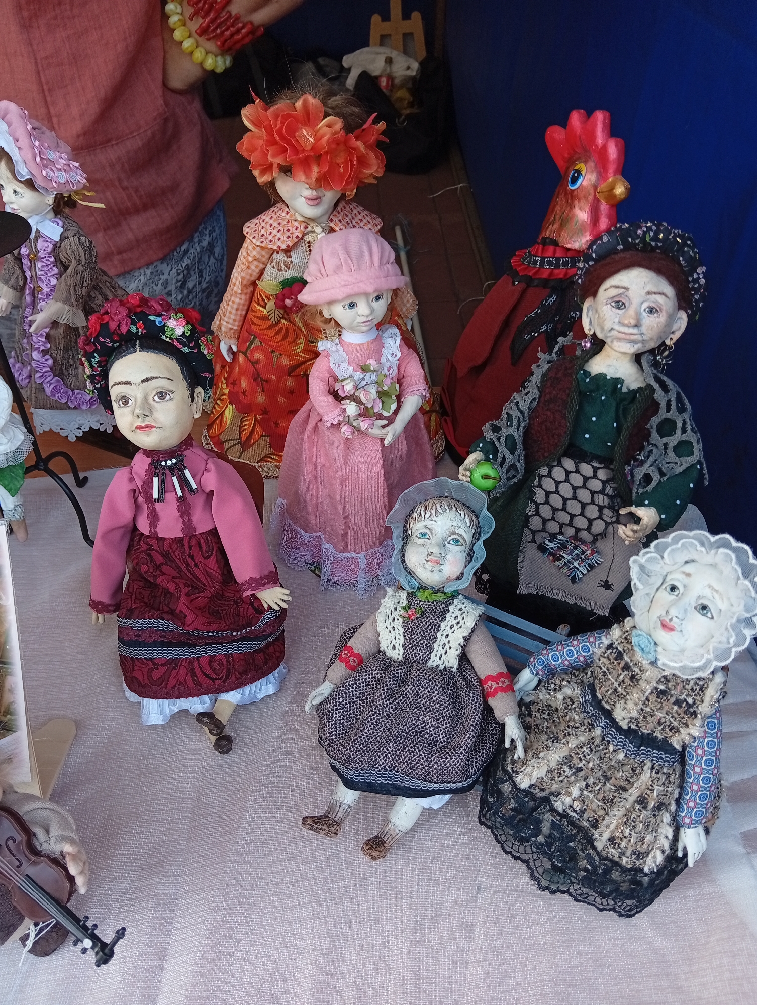 The image depicts a collection of handmade dolls arranged on a pink cloth surface, likely a market stall. In the background, a person wearing a red-brown shirt and a blue patterned skirt is visible. To the right, a large doll with a red rooster headdress stands. Several other dolls are present, including one dressed in traditional clothing resembling Frida Kahlo, a doll in a pink dress holding flowers, a doll in a brown checkered dress, and two elderly-looking dolls with ruffled bonnets. A miniature violin rests on the left side of the image. The lighting suggests an outdoor setting, possibly a street market or fair. The specific location in Kostanay, Kazakhstan, is not discernible from the image itself; however, the presence of traditional-looking dolls and a vibrant display suggests a local artisan market or craft fair, common in such cities.