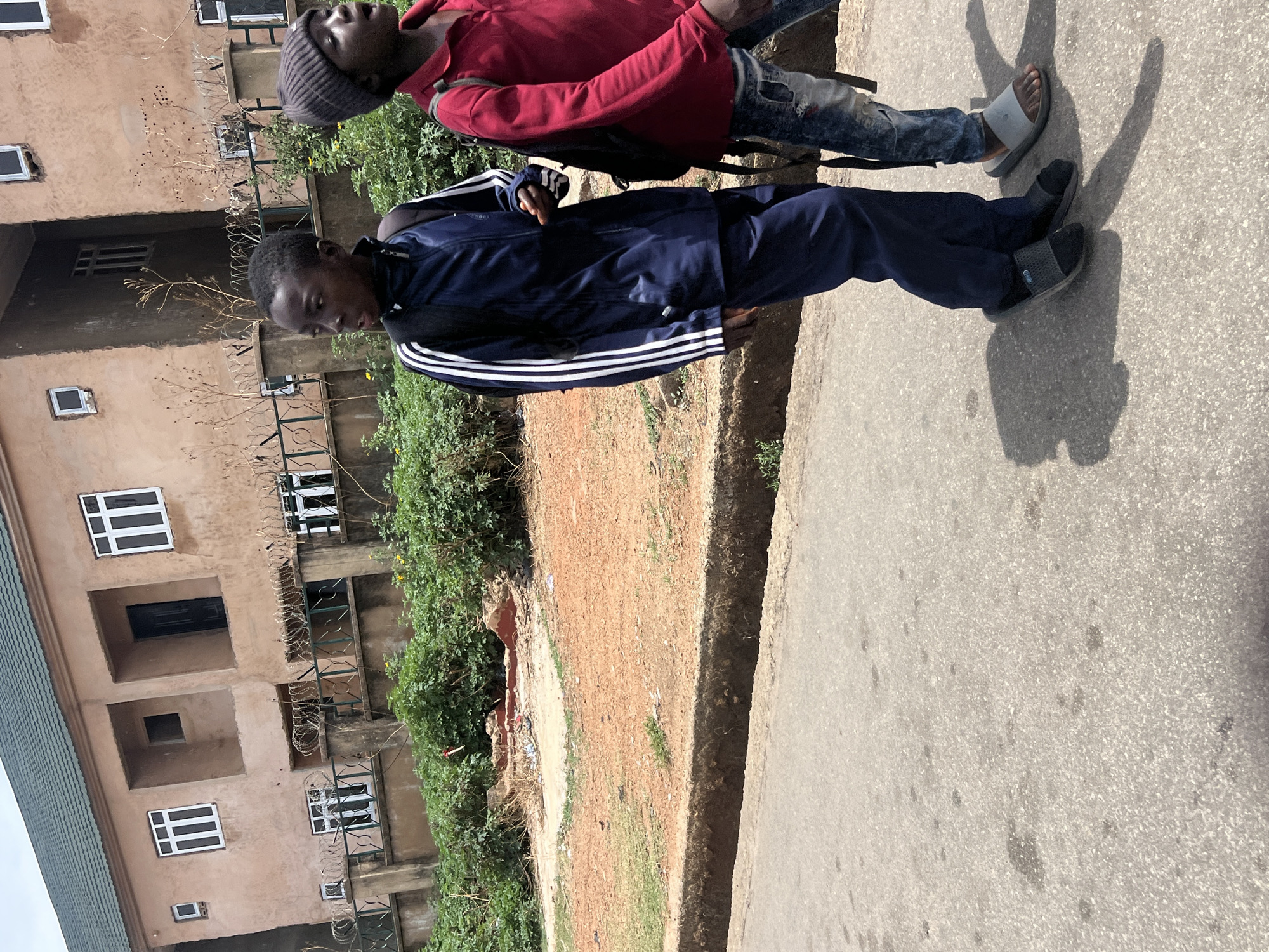 The image, rotated ninety degrees clockwise, captures two young males standing outdoors on what appears to be a paved path or road in Jos, Nigeria.
The subject in the foreground wears a dark blue full-zip tracksuit with white triple stripes on the sleeves, indicative of Adidas branding. He also wears dark slippers. His expression is neutral, looking slightly downwards and to the side.
Behind him, another young male is visible, wearing a red hooded sweatshirt, a grey beanie, and light blue distressed jeans. He has light-colored slide sandals on his feet. He is looking upwards and slightly to the right, with a neutral expression. Both individuals cast distinct shadows on the ground, suggesting bright sunlight.
The setting includes a grey asphalt surface where the subjects are standing. To their left, there is a patch of reddish-brown dirt and green foliage. In the background, a multi-story building with a light brown or sandy-colored facade is visible. The building features several rectangular windows with white frames and black grilles. Multiple balconies or window openings are secured with black metal fences, some of which are topped with barbed wire. A green-tiled roof is partially visible on the left side of the frame. The overall impression is an urban or semi-urban street scene under clear, sunny daylight.