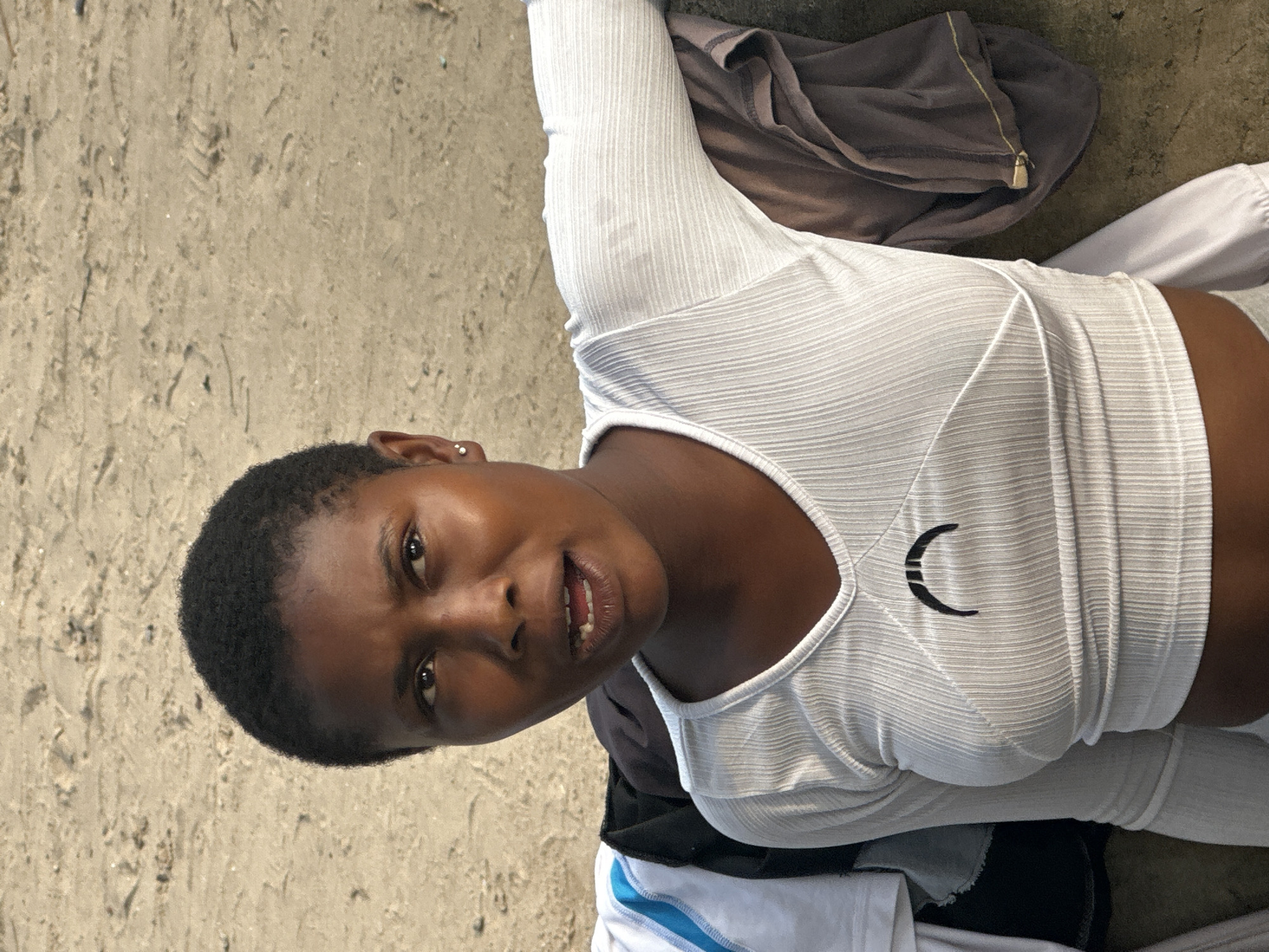 A young Black woman with short, dark hair and small stud earrings is seated outdoors, leaning against a textured, sandy surface. She is wearing a white, long-sleeved, ribbed crop top with a V-neck and a black crescent moon design centered on the chest. To her left, there are several pieces of clothing, including a white t-shirt with blue and white trim, and a dark jacket. A brown garment is draped over the surface behind her right arm. The background is a plain, sandy area. The woman is looking slightly to her right and appears to be speaking, with her mouth open and teeth visible. The lighting suggests it is daytime.