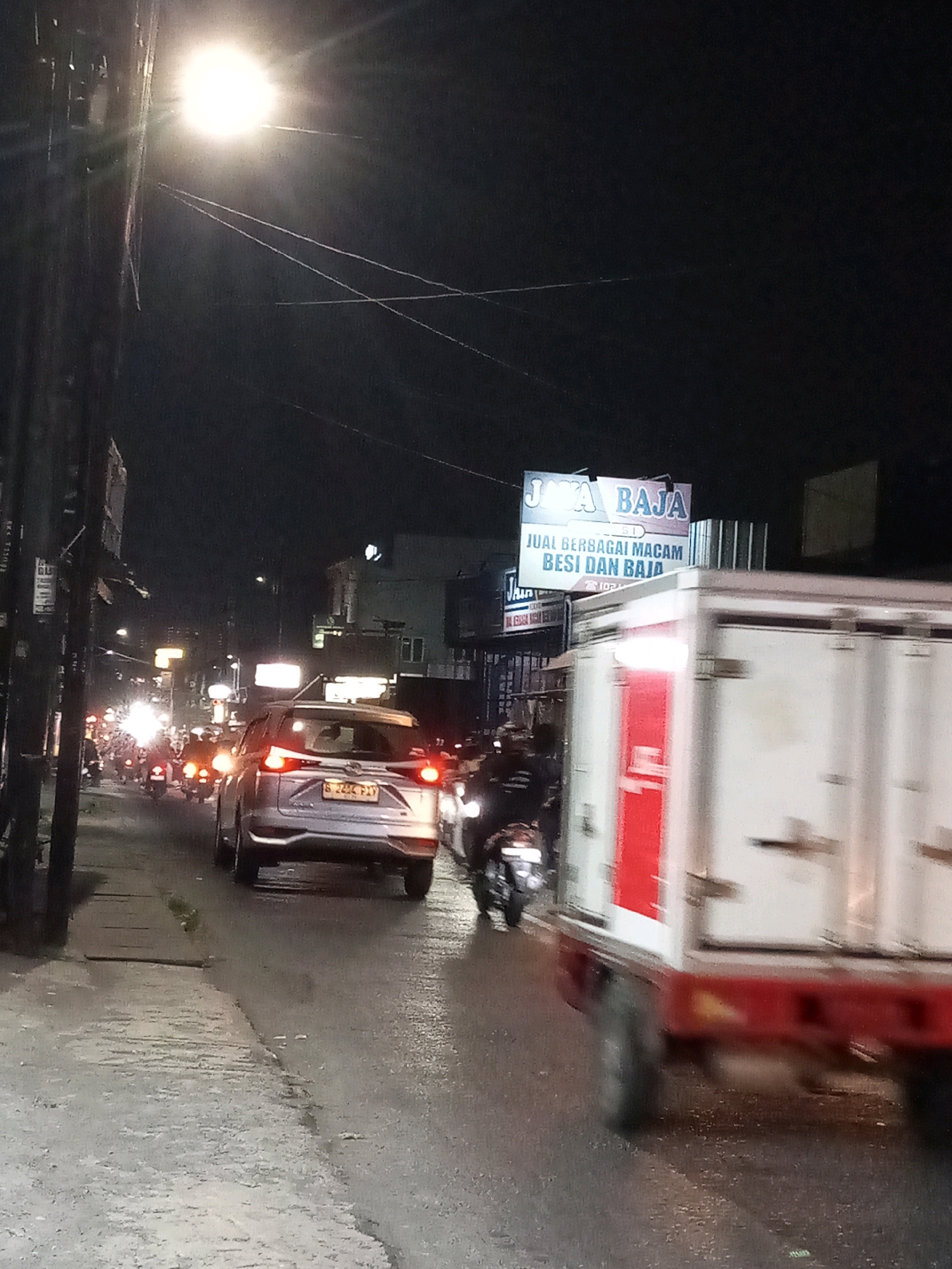 A nighttime, low-angle shot captures a busy street in Bekasi, Indonesia, likely a commercial area. A bright streetlamp illuminates the left side of the frame, casting long shadows. The road is wet, indicating recent rain or a damp environment. Traffic is moderate, with several motorcycles and a silver SUV visible. The SUV, a 2023 or newer model, has its brake lights illuminated. A white box truck, heavily blurred due to motion, is in the foreground, obscuring a significant portion of the right side of the image. On the left side of the road, a utility pole with wires runs vertically. A sign for "JAVA BAJA" with text indicating "JUAL BERBAGAI MACAM BESAI DAN BAJA" (selling various types of iron and steel) is mounted on a building in the background. The overall atmosphere is one of a bustling urban street at night, with the characteristic signs and traffic of Indonesia.