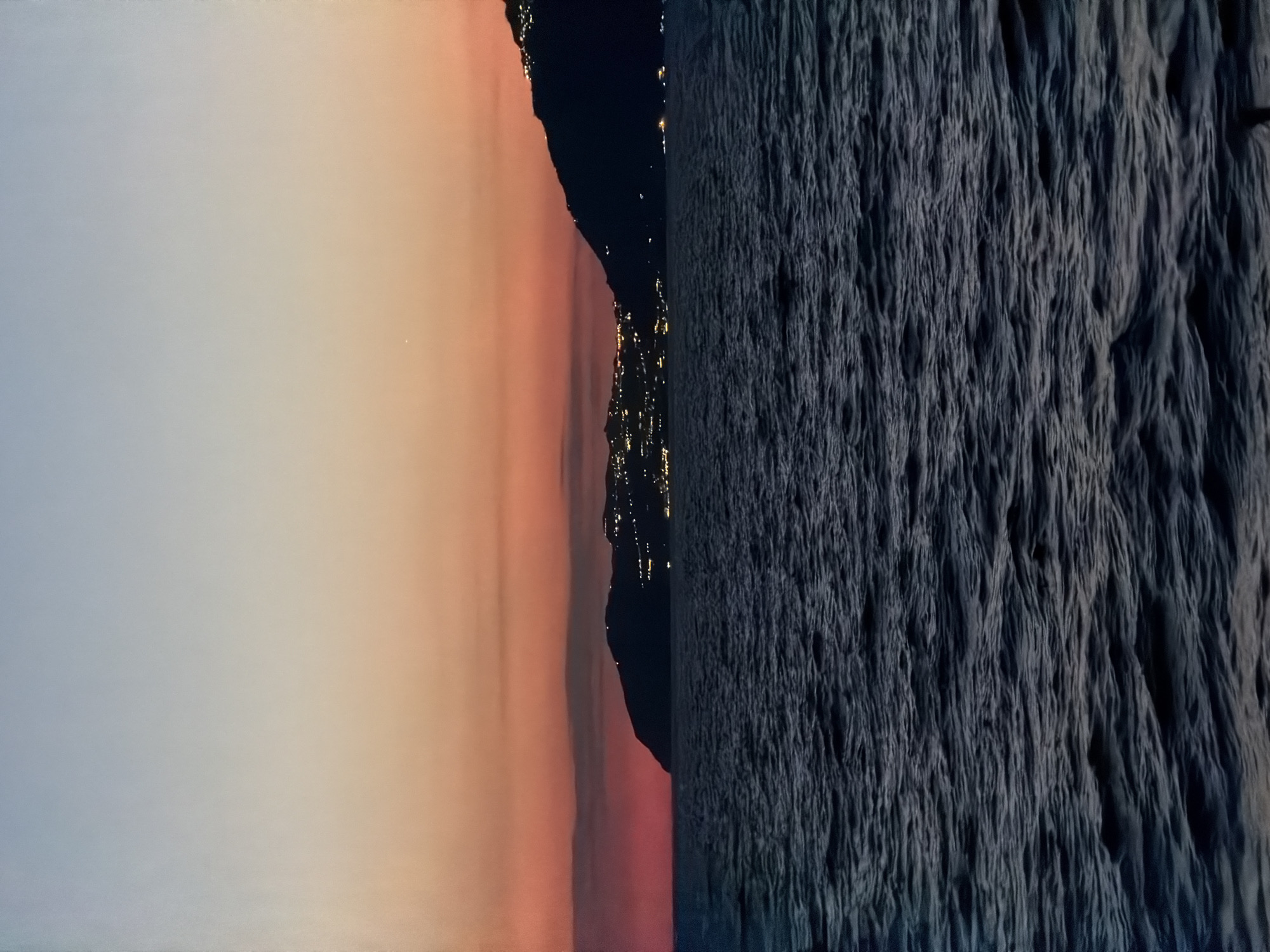 The image shows a rotated view of Ischia, Italy. On the left, a pale sky transitions to a warm, peachy hue near the ground. In the middle, a dark landmass with scattered yellow lights resembling a city at night extends upwards. To the right, a textured, dark surface resembling rock or vegetation occupies a significant portion of the frame.