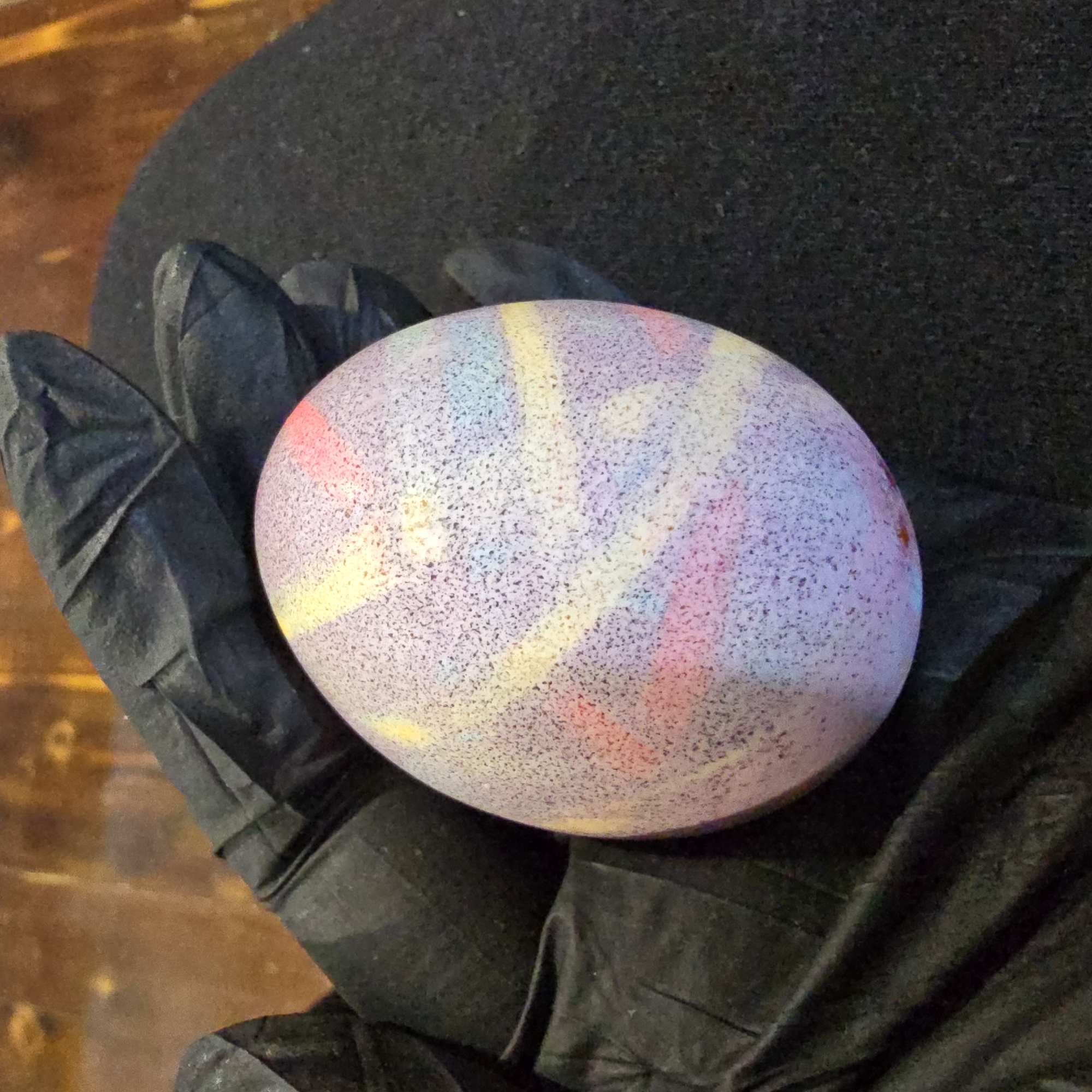 A gloved hand holds an Easter egg. The egg is primarily lavender with spots and light pastel colored streaks. The glove is black. The background is a table. The location of the photo is unknown.