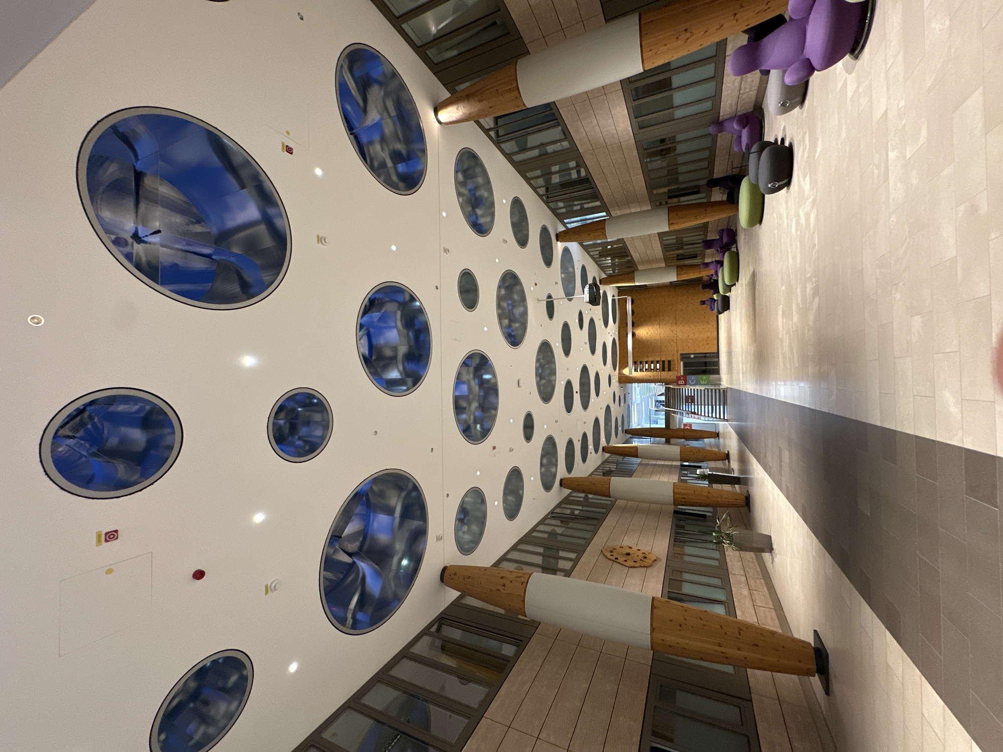 The image shows a wide, modern hallway with a high, white ceiling featuring numerous circular and oval openings that reflect the interior lighting and the ceiling structure. The walls are lined with floor-to-ceiling windows and paneling that resembles light-colored wood. Large, cylindrical columns, covered in sections of white material and natural wood, support the ceiling. The floor is tiled in a light neutral color, with a darker tiled strip running down the center of the hallway. In the distance, on the right side of the hallway, there are several colorful, bulbous armchairs and poufs, suggesting a waiting or lounge area. A staircase is visible further down the hallway. The overall impression is one of a contemporary and spacious interior, likely a public building or office space. There are no people visible in the image. The image is not located in Garmisch-Partenkirchen, Germany.