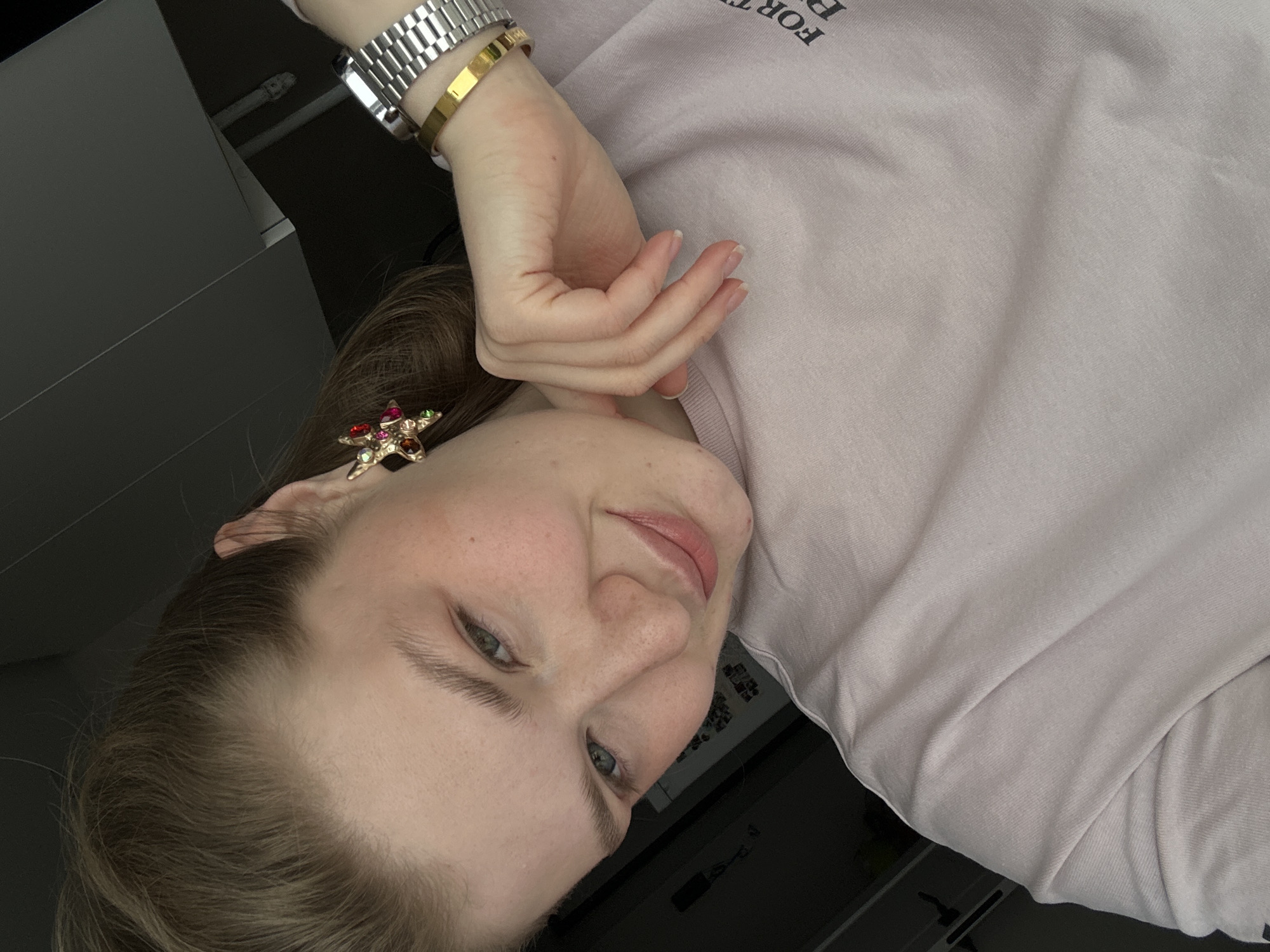 The image shows a woman with light brown hair and fair skin, wearing a light pink sweatshirt and jewelry. She is wearing a silver watch, a gold bracelet, and a star-shaped earring with colorful stones. She is posing with her hand near her face. The background appears to be a blurred interior space with dark elements, possibly furniture or other objects. The image does not provide any location information or evidence that it was taken in Kazan, Russia.