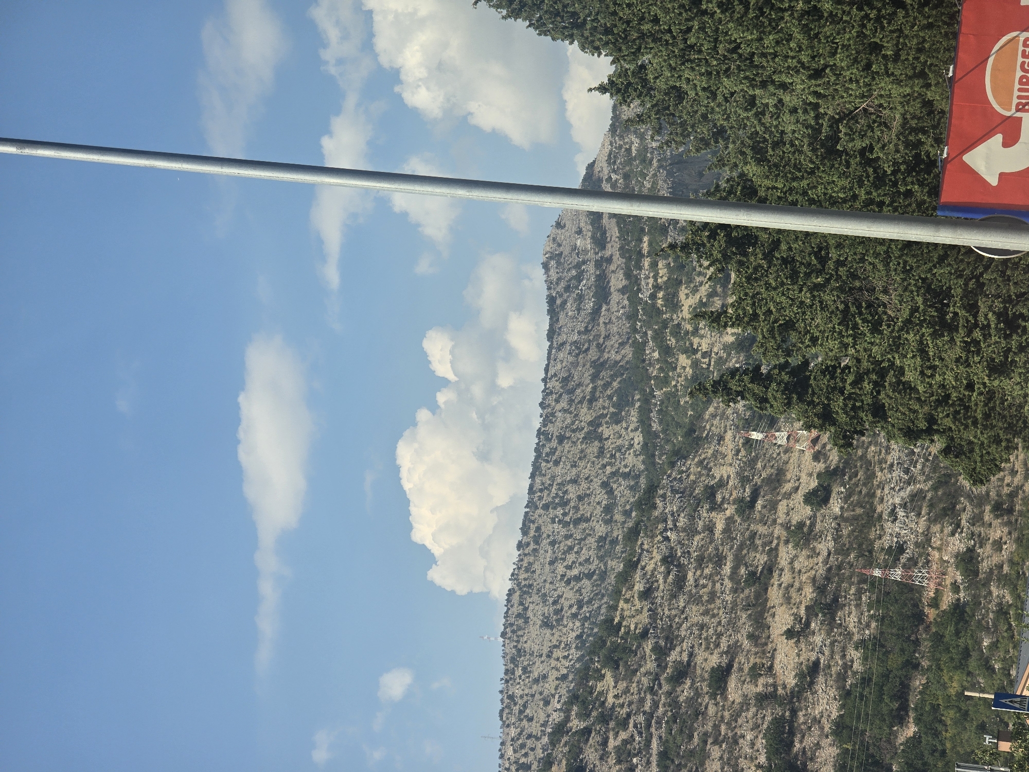 The image depicts a mountainous landscape under a blue sky with scattered white clouds, likely taken from a street-level perspective in Podgorica, Montenegro. A prominent silver pole runs vertically through the right side of the frame, obscuring a portion of the view. In the foreground, on the right, a portion of a red billboard with a white arrow and the "BURGER KING" logo is visible. Behind the billboard and pole, a dense line of dark green coniferous trees forms a natural barrier.
Beyond the trees, a rugged, rocky mountainside dominates the middle ground. The terrain is sparsely vegetated with small trees and shrubs. Two lattice towers, one red and white and the other white, are visible on the mountainside, suggesting the presence of communication or power lines. The background consists of more distant, hazy mountains under the bright sky. The overall scene conveys a sense of a semi-urban environment bordering a natural, mountainous region.