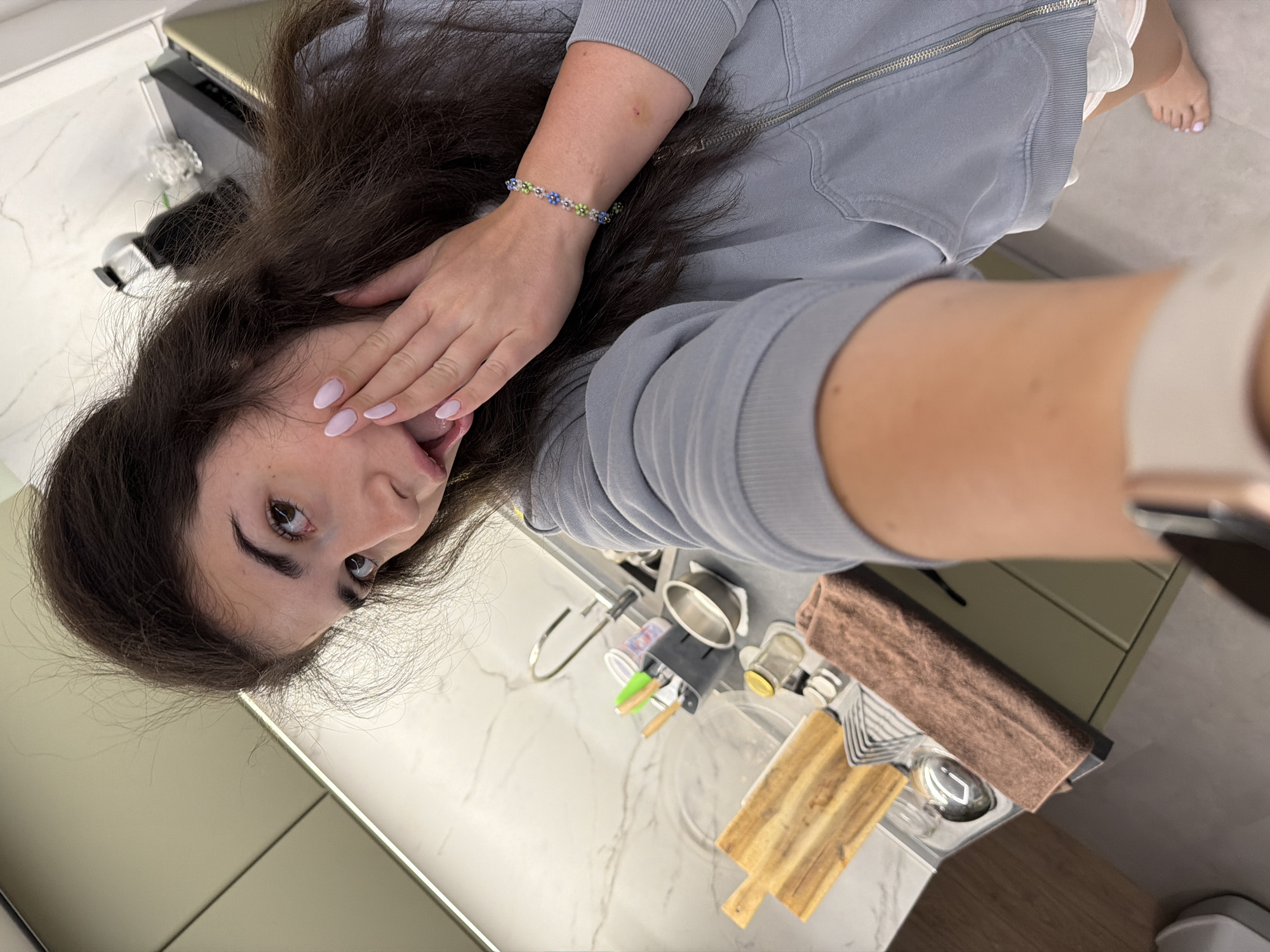 A young woman with long, dark hair is taking a selfie in a modern kitchen. She is wearing a light blue zip-up hoodie and white shorts. Her left hand is placed on her cheek, and her mouth is open. She has light pink nail polish on her fingernails and is wearing a beaded bracelet on her left wrist. The kitchen features a marble backsplash, a sink with a faucet, a cutting board, and various kitchen utensils. The overall impression is informal and domestic. The image does not contain any specific information indicating the location in Sofia, Bulgaria.