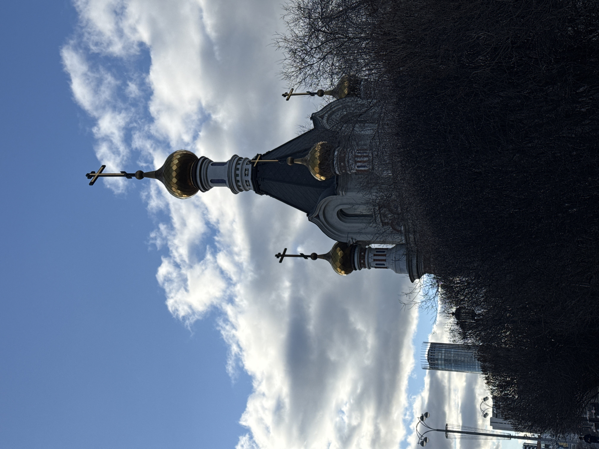 The image shows a Russian Orthodox church, likely located in Yekaterinburg, Russia. The church has a traditional design with multiple golden onion domes topped with crosses. The domes and the structure are partially obscured by trees, possibly bare branches, suggesting it may be winter or early spring. The sky is visible with a mix of blue sky and white clouds. A tall building is visible in the background.
