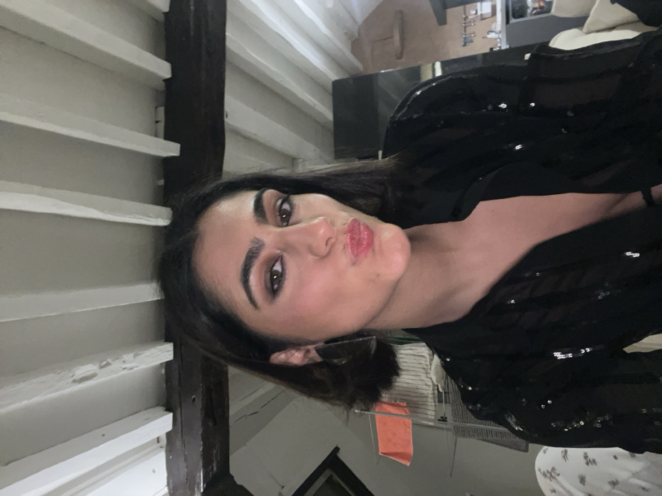 A woman with dark hair and light skin poses indoors with a slight pout. She wears a black top with embellishments. The background includes white slat paneling and a dark beam, creating a rustic ambiance. Behind her, a glimpse of a room with a black surface and a possible window with a towel hanging nearby is visible. The setting suggests a cozy interior, possibly a home.