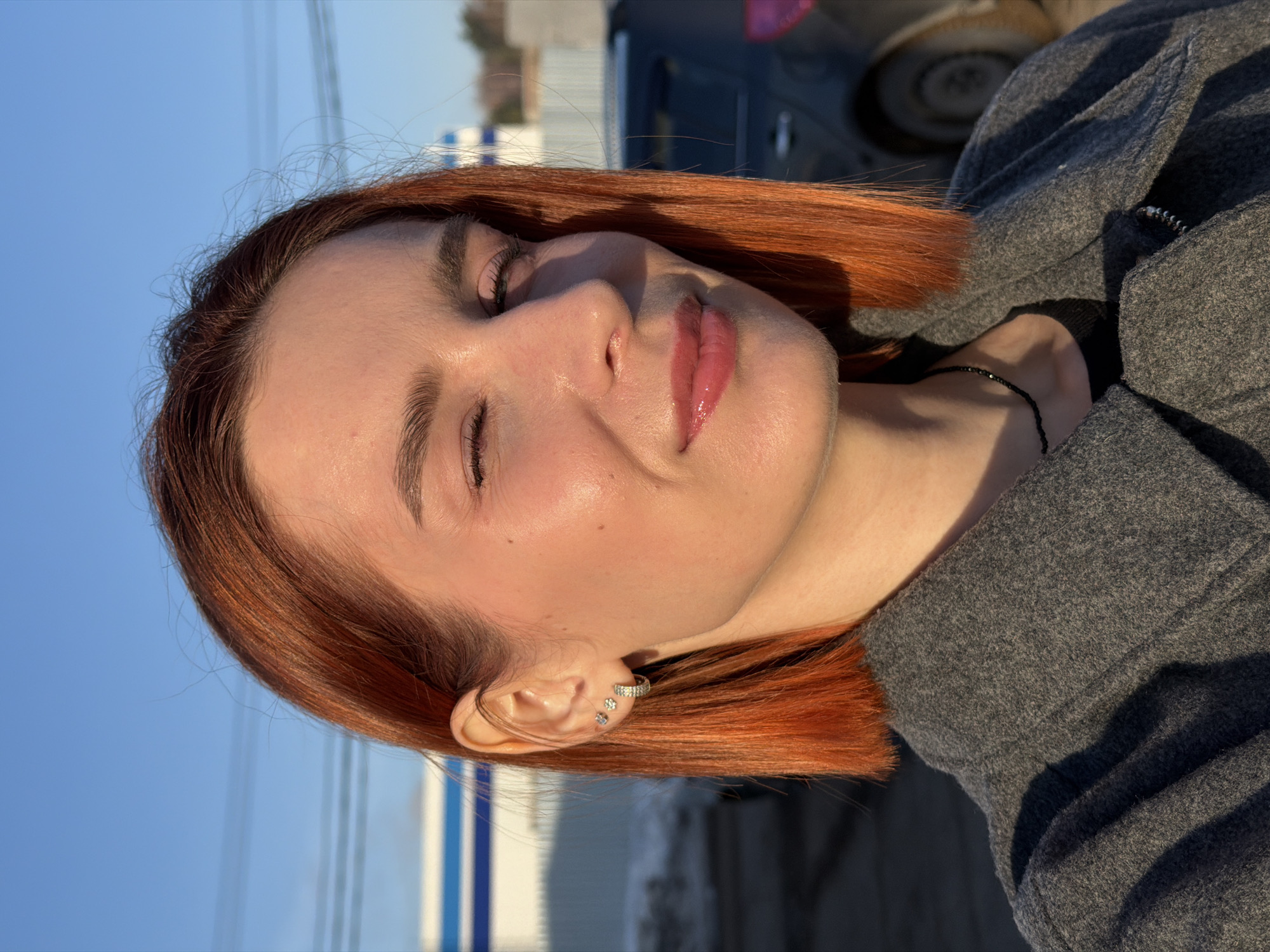 A medium close-up of a woman with auburn hair and fair skin with her eyes closed. She is wearing multiple earrings in her left ear and a gray jacket. The background is a bright blue sky on the left side and a partial view of an out-of-focus vehicle on the right side, along with a white wall with blue accents. The photo was taken outdoors. The image lacks clear indicators of location in Chaikovsky, Russia.