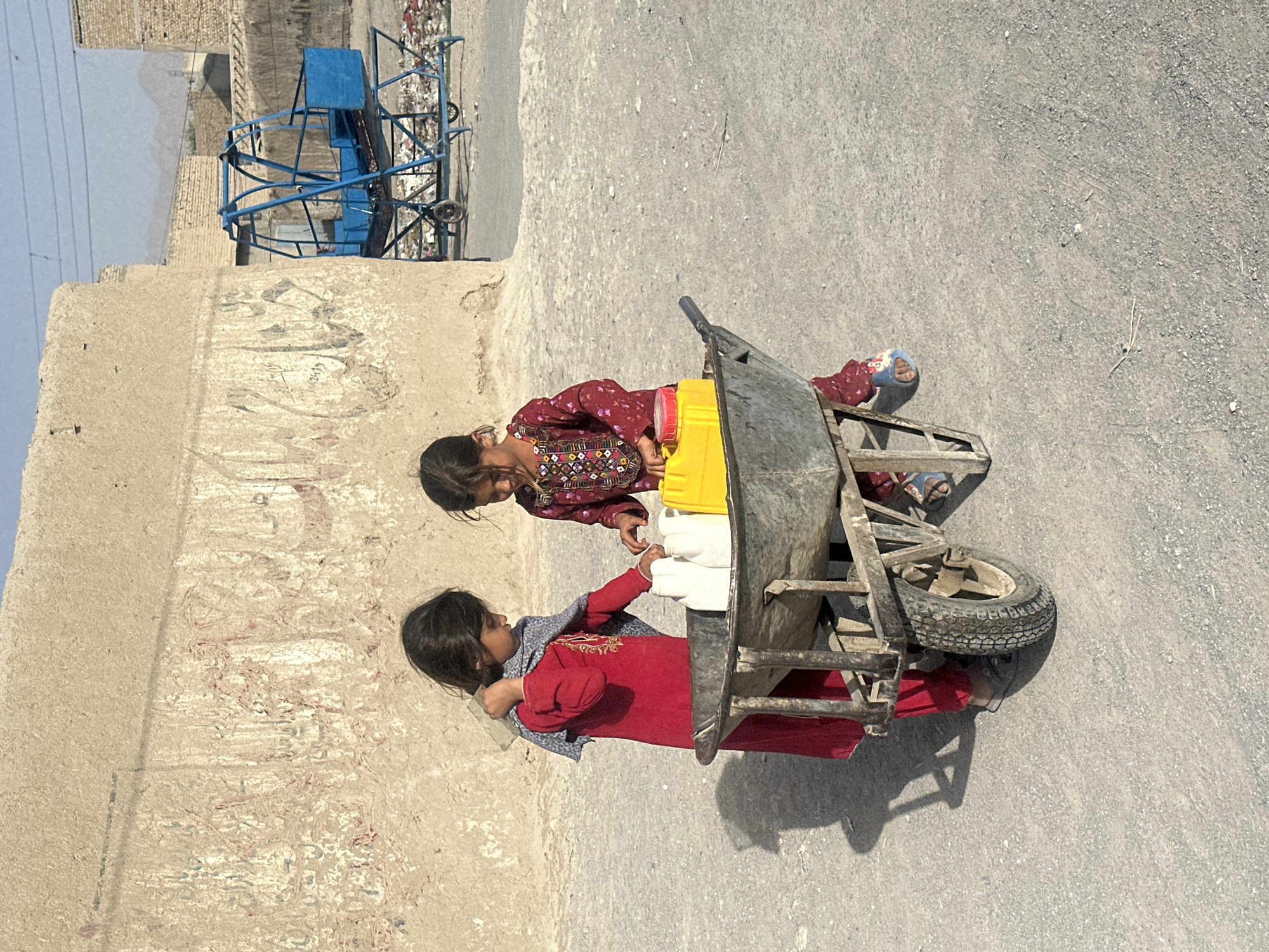 Two young girls are in a dusty, outdoor environment. The girl on the left wears a red dress and a grey scarf, and she is carrying a white container on her shoulder. She is standing next to a wheelbarrow. The girl on the right wears a red, patterned dress and is reaching into the wheelbarrow. The wheelbarrow contains two white containers and a yellow container. The background shows a textured, light-colored wall with some faded markings, and further in the background, there is a blue metal structure and buildings. The ground is a rough, grey surface.