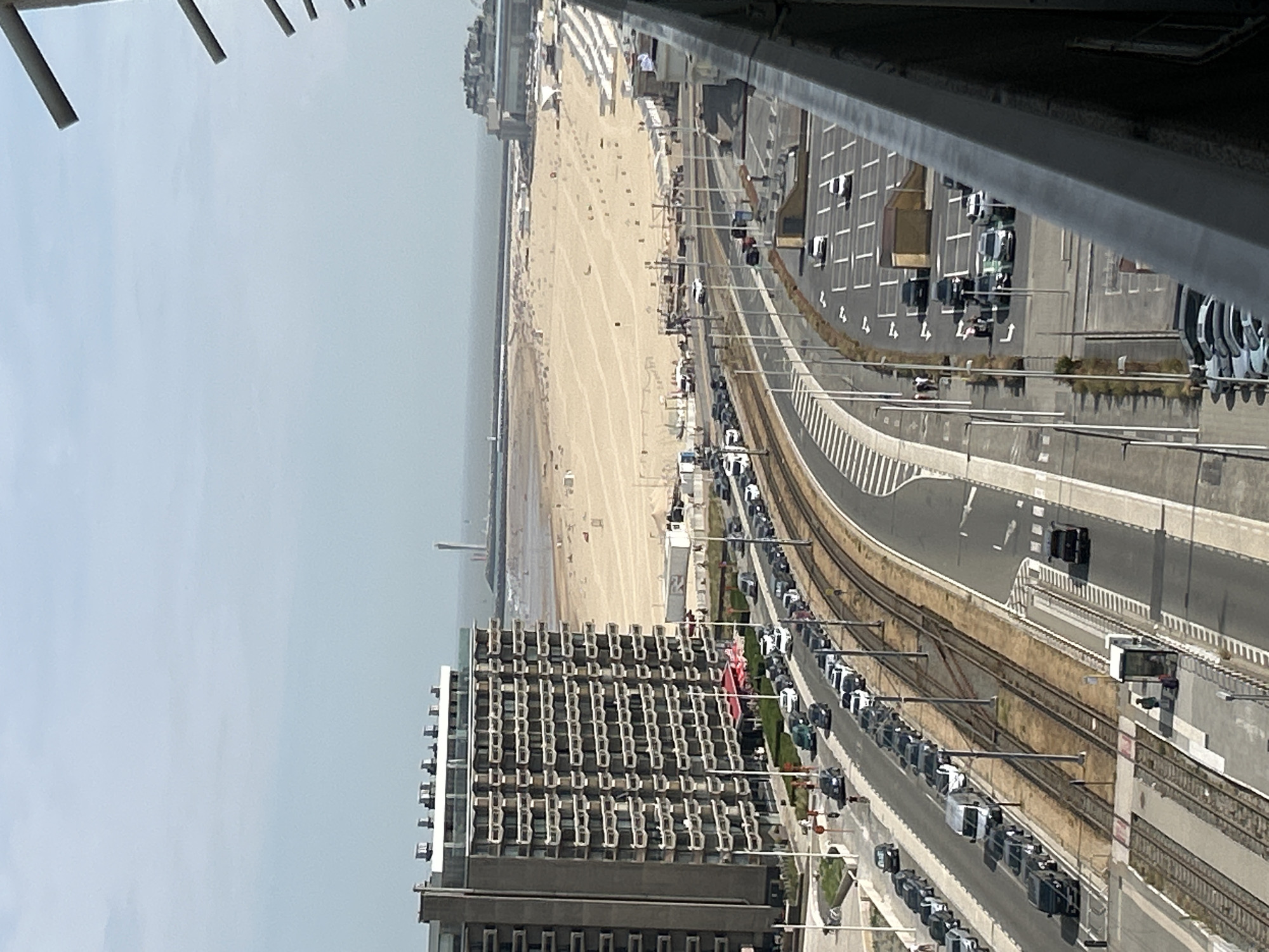 The image captures a view of the beach and coastline in Ostend, Belgium, on a hazy day. In the foreground, a multi-story building with a distinctive facade of balconies dominates the left side of the frame. Adjacent to the building, a road runs parallel to train tracks, with numerous cars parked along the side and in a parking lot. Further in the distance, a wide sandy beach stretches along the coastline, dotted with beach chairs and umbrellas. The sea is visible beyond the beach, meeting the horizon under a pale sky. A lighthouse or tower stands on the far left of the horizon, and another distant building structure is visible on the right. There are people visible on the beach and near the road, and a single person stands on a train platform. The overall scene depicts a seaside urban environment.