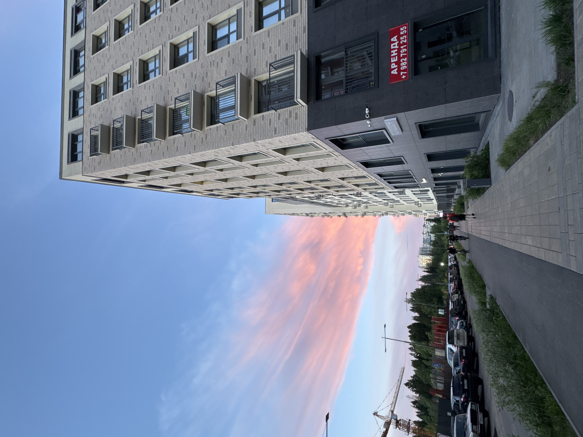 The image depicts a multi-story building on a street in Izhevsk, Russia. The building is constructed with brick and grey stone, with a visible "Аренда" (Rental) sign and a phone number. The sky shows pink clouds, indicating either sunset or sunrise. The street features a sidewalk, a row of parked cars, trees, and streetlights. People are seen walking on the sidewalk. A construction crane is visible in the background.