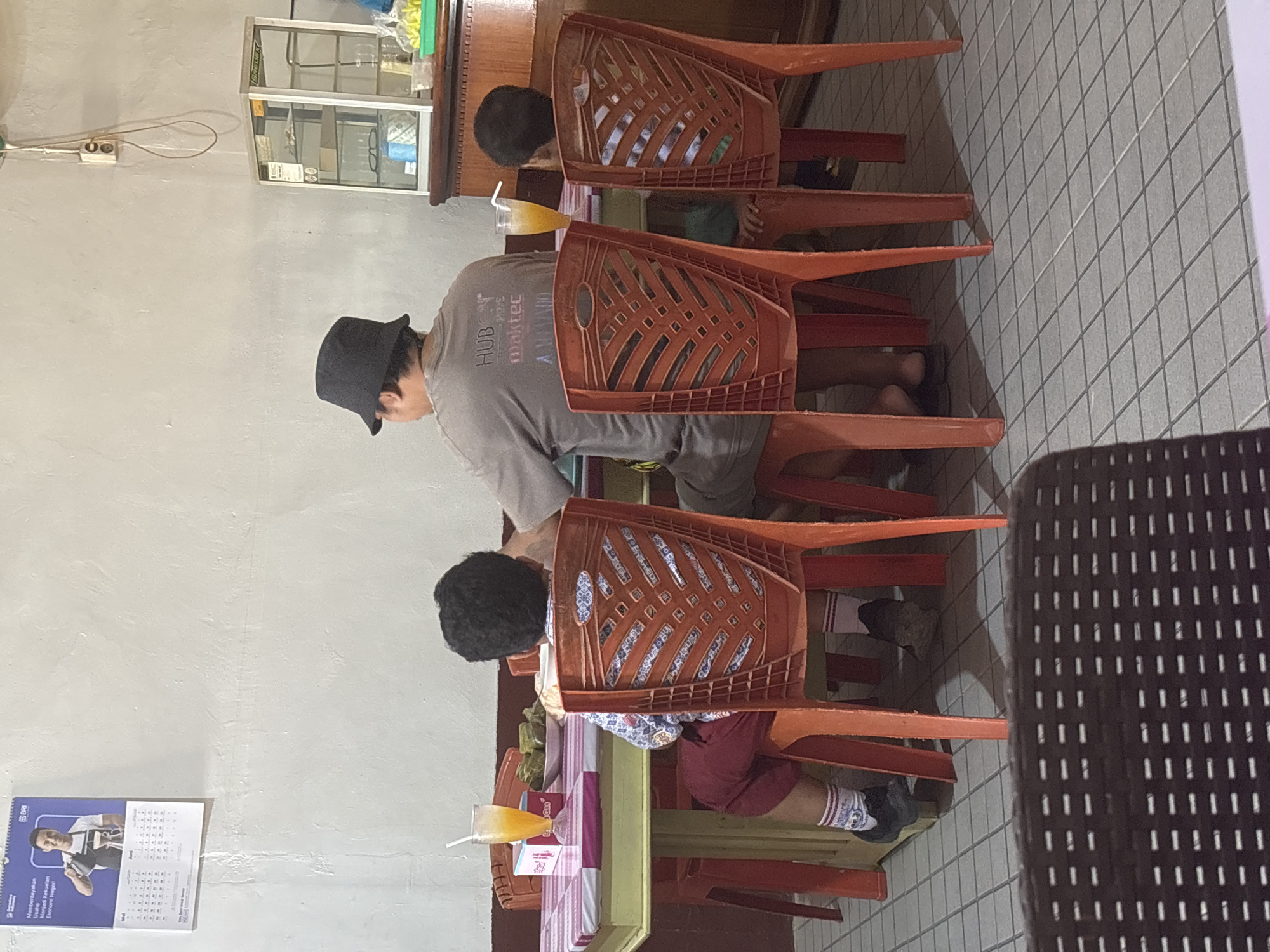 The image shows a group of people, likely a family, sitting at a table in an indoor setting. A man wearing a black bucket hat is seated on the right, facing away from the camera. He is wearing a grey t-shirt with text that appears to say "HUB maktec". Two children are seated in front of him, also facing away from the camera. One child on the left is wearing a patterned shirt and red shorts, with white socks and dark shoes. The other child, on the right, is partially visible. A glass of orange juice with a straw is on the table in front of the child on the left, along with a plate of food that appears to be wrapped in leaves. The table has a pink and white checkered tablecloth. The chairs are orange plastic. In the background, a calendar is hanging on the wall, depicting a man pouring coffee. To the right, a glass display cabinet is visible, containing various items including what appears to be glasses. The floor is tiled with a grid pattern. The environment appears to be a casual dining establishment or a home dining area. The scene is devoid of any specific indicators that it is located in Merauke, Indonesia.