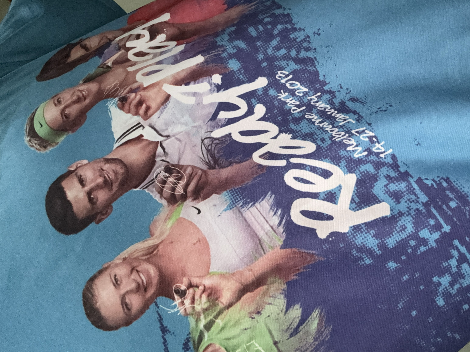 A close-up, slightly angled shot of a light blue t-shirt features a graphic print of four tennis players and text. From left to right, the players are Maria Sharapova, Novak Djokovic, and two others, partially visible. They appear to be holding pens and signing something. The graphic also includes the text "Ready?" in large white script, with "Melbourne Park 14-27 January 2013" written below it. The background of the graphic is a distressed blue and teal pattern. The t-shirt is laid flat or displayed against an unseen surface, with the fabric showing folds and texture. The image is focused on the t-shirt's design. The location of Haizhu District, China is not discernible from the image.