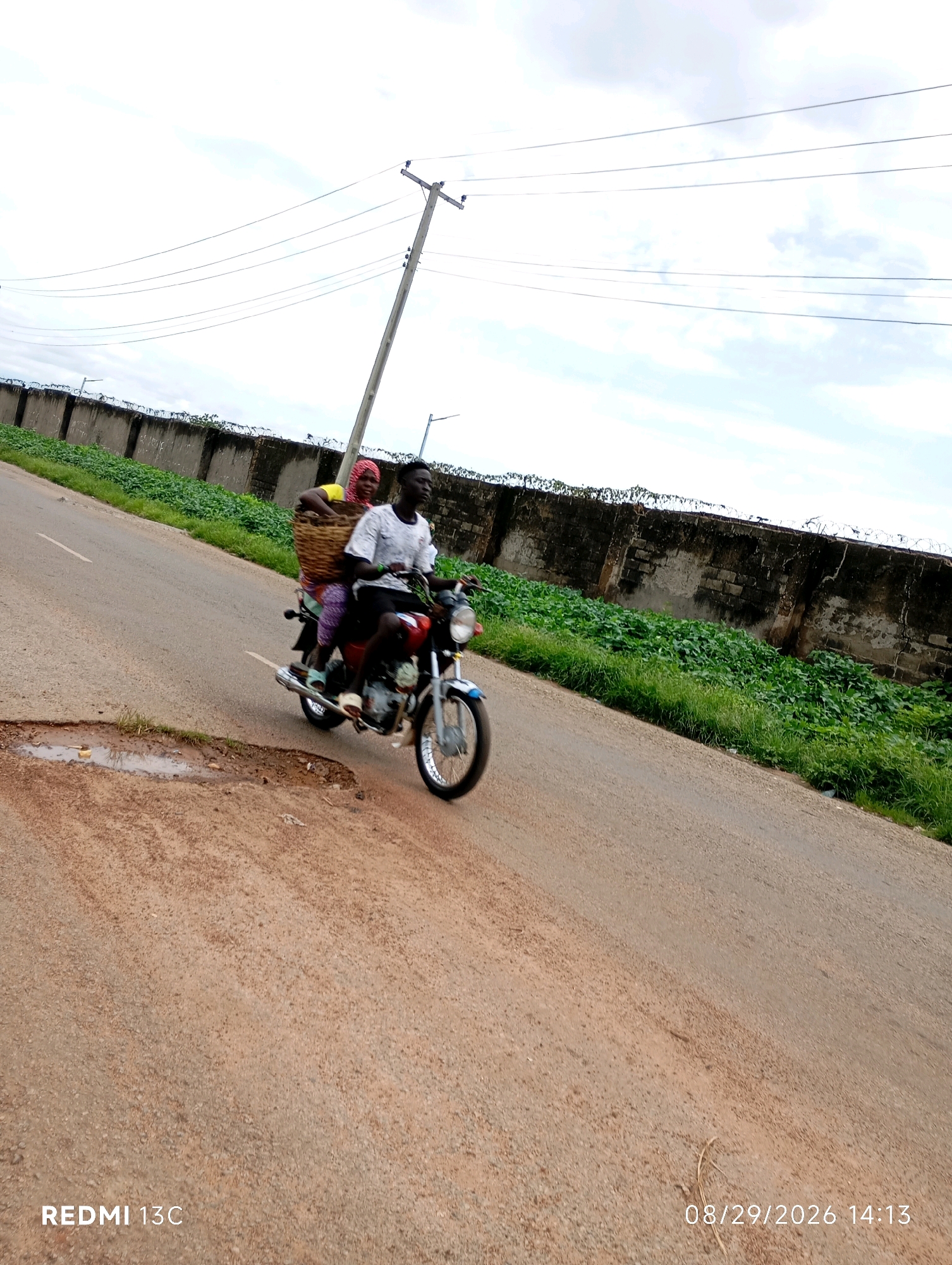 In Shendam, Nigeria, two individuals are traveling on a motorcycle. The person in the rear, a woman wearing a yellow top and a headscarf, carries a large woven basket filled with goods. The person in front, a man wearing a white t-shirt with a graphic design and dark trousers, is operating the motorcycle. They are riding on a paved road with a sandy shoulder. A large, dark brick wall with barbed wire runs along the right side of the road, with lush green vegetation growing in front of it. A utility pole with electrical wires stands further down the road. The sky above is overcast with scattered clouds. The timestamp "REDMI 13C" and "08/29/2026 14:13" are visible in the lower portion of the image.