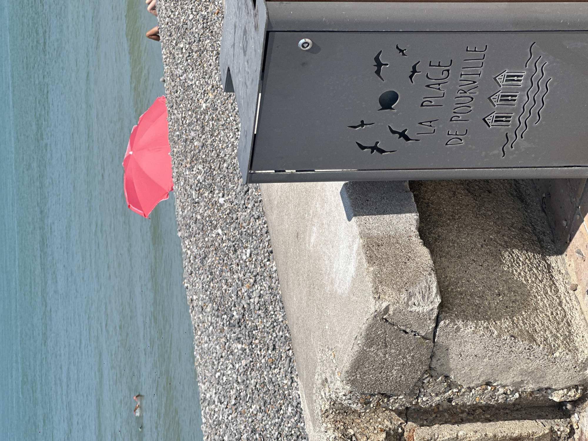 This image depicts a view of the pebble beach at Pourville, a seaside resort located within the commune of Hautot-sur-Mer, France. In the foreground, a dark gray metal bin with decorative cutouts of birds, a fish, and beach huts is positioned on a concrete ledge. The text "LA PLAGE DE POURVILLE" is also cut into the bin. The concrete ledge shows signs of age and wear, with cracks and exposed aggregate. Behind the bin and ledge, a stretch of grey pebbles forms the beach, leading to the ocean. The water is a clear blue-green. In the middle ground, a person is swimming in the sea. Further out, a bright red umbrella is visible on the beach, with the legs of at least one person visible near it. The sky is not visible in this frame.