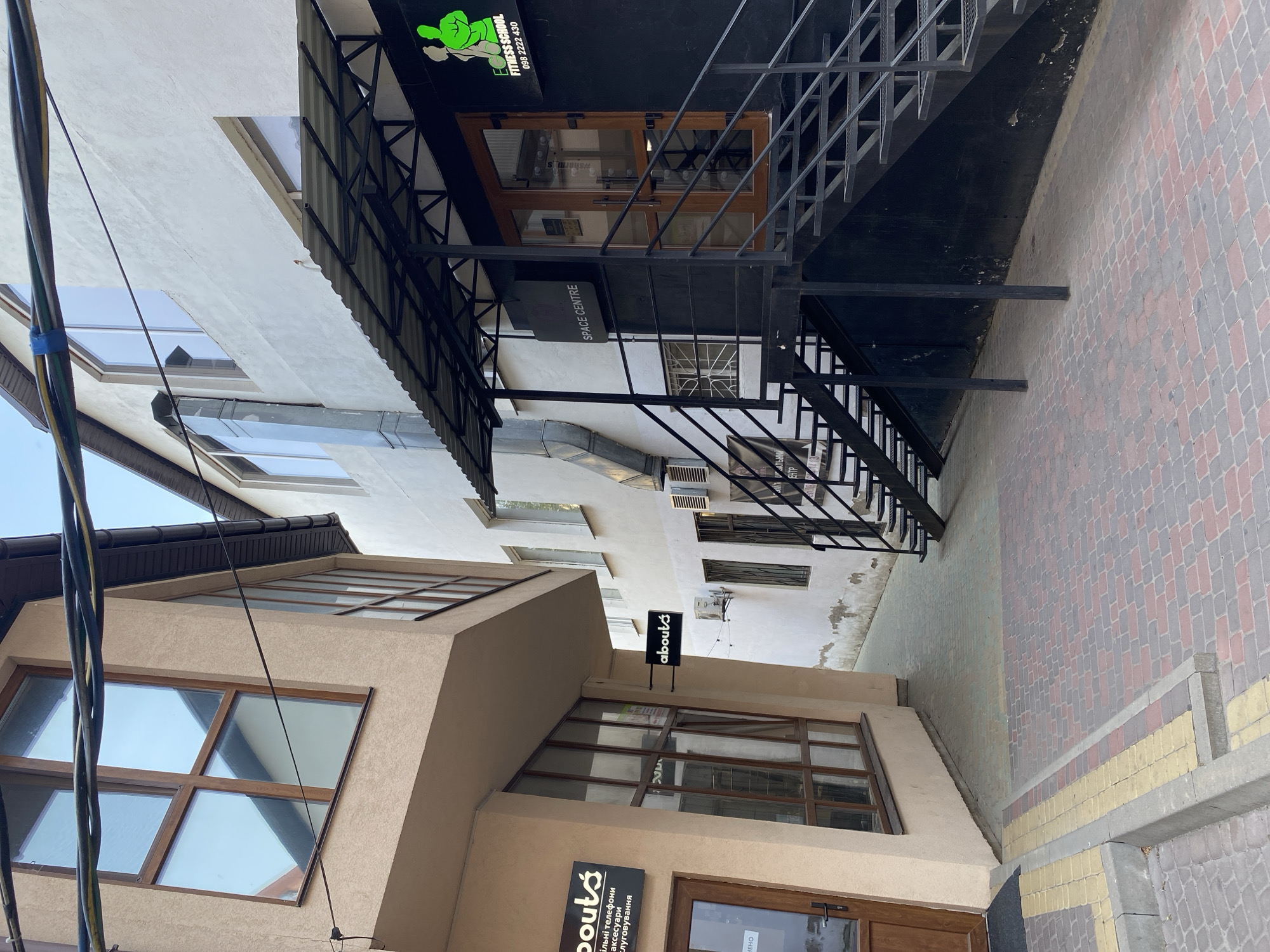 The image captures a narrow alleyway between two buildings in Vinnytsia, Ukraine. On the left, a light beige building features large windows with wooden frames, and a dark sign with white text reads "aboutś" and other Ukrainian text. A glass door is visible on the ground floor.
On the right, a white building has multiple windows. A black awning extends over a set of dark metal stairs leading up to an entrance. The entrance is marked by a black facade with a prominent neon green logo of a figure performing a squat. Below the logo, white text reads "FITNESS SCHOOL" and a phone number. A sign with "SPACE CENTRE" in white text on a black background is also visible on this building.
The alleyway is paved with interlocking bricks in a pattern of red and grey. The sky is visible at the top of the image, with power lines crossing overhead. The overall scene depicts an urban commercial area. There are no people visible in the image.