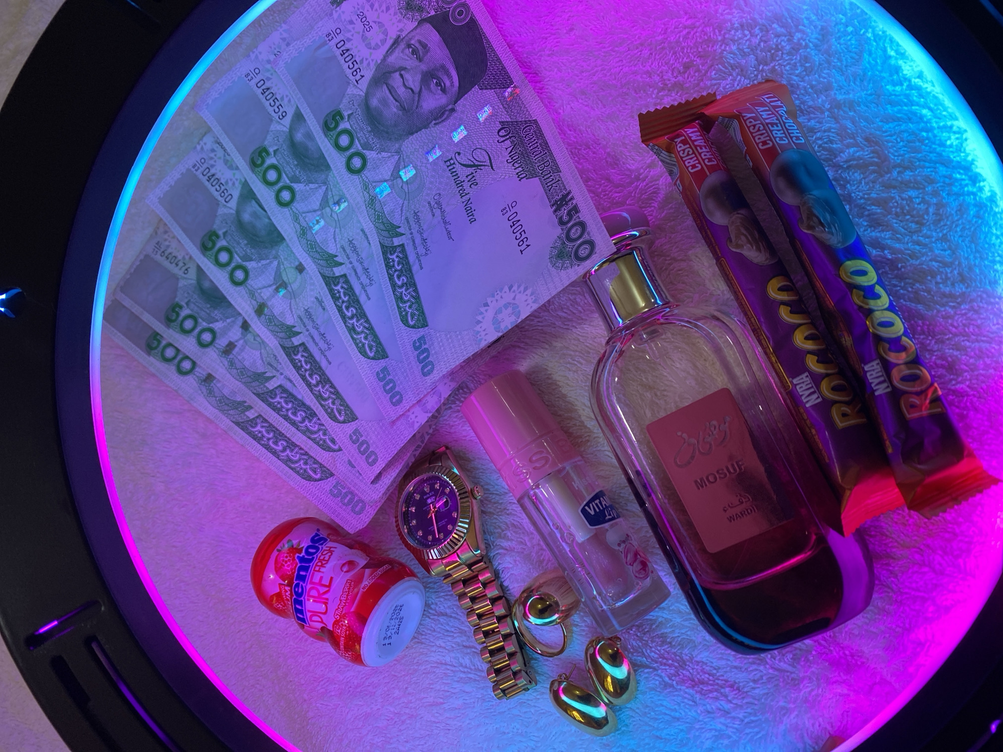 The image displays a flat lay arrangement of various personal items and consumables, resting on a white, textured fabric surface and illuminated by a ring light casting vibrant blue and pink hues.
The central subjects include several 500 Nigerian Naira banknotes, clearly showing the portrait of Dr. Nnamdi Azikiwe. Visible text on the currency includes "Central Bank Of Nigeria," "Five Hundred Naira," "N500," and serial numbers such as Q 040561, Q 040559, Q 040560, and AL 73 640476. Signatures of the Governor and Director of Currency Operations are also discernible, along with the year "2025."
Other items in the arrangement are a bottle of "MOSUF Wardi" perfume and a smaller clear bottle with a pink cap, which appears to be a cosmetic product, with "VITA" and "Lip" partially visible on its label. Accompanying these are a gold-toned watch with "ROLEX" visible on its face, and two gold-toned rings.
Consumables include a red container of "mentos PURE FRESH STRAWBERRY FLAVOUR," marked "NO ADDED SUGAR" and showing expiry dates 13/06/2025 and 09/12/2026. Two "ROCOCO NYRA CRISPY CREAMY CHOCOLATI" bars are also present.
The scene appears to be a curated display, possibly for an aesthetic presentation or advertisement. The items are arranged neatly, highlighting their details under the colorful light. The presence of Nigerian Naira notes places the context in Nigeria, specifically Maigizo as indicated. No activity is taking place; it is a still life composition.