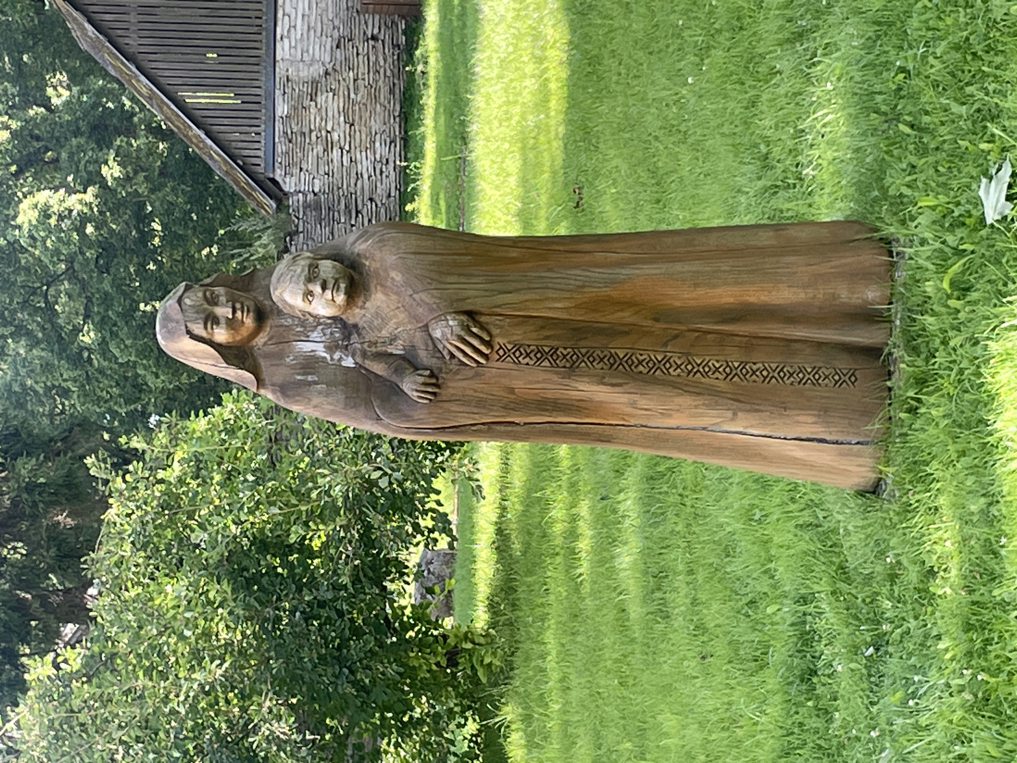 A full-length, upright wooden sculpture of a woman embracing a child stands in a grassy outdoor area. The figures are carved from a single piece of wood, with the woman's head covered by a hood. The woman's hands rest on the child, who is positioned in front of her and partially enveloped by her robe. A decorative band of geometric patterns is carved down the front of the woman's robe. The sculpture is positioned in the foreground, with a blurred background of green trees, a stone building, and a wooden structure. The ground is covered in lush green grass, with some small white flowers scattered throughout. The scene appears to be a tranquil park or garden setting. This sculpture is located in Jõelähtme, Estonia.