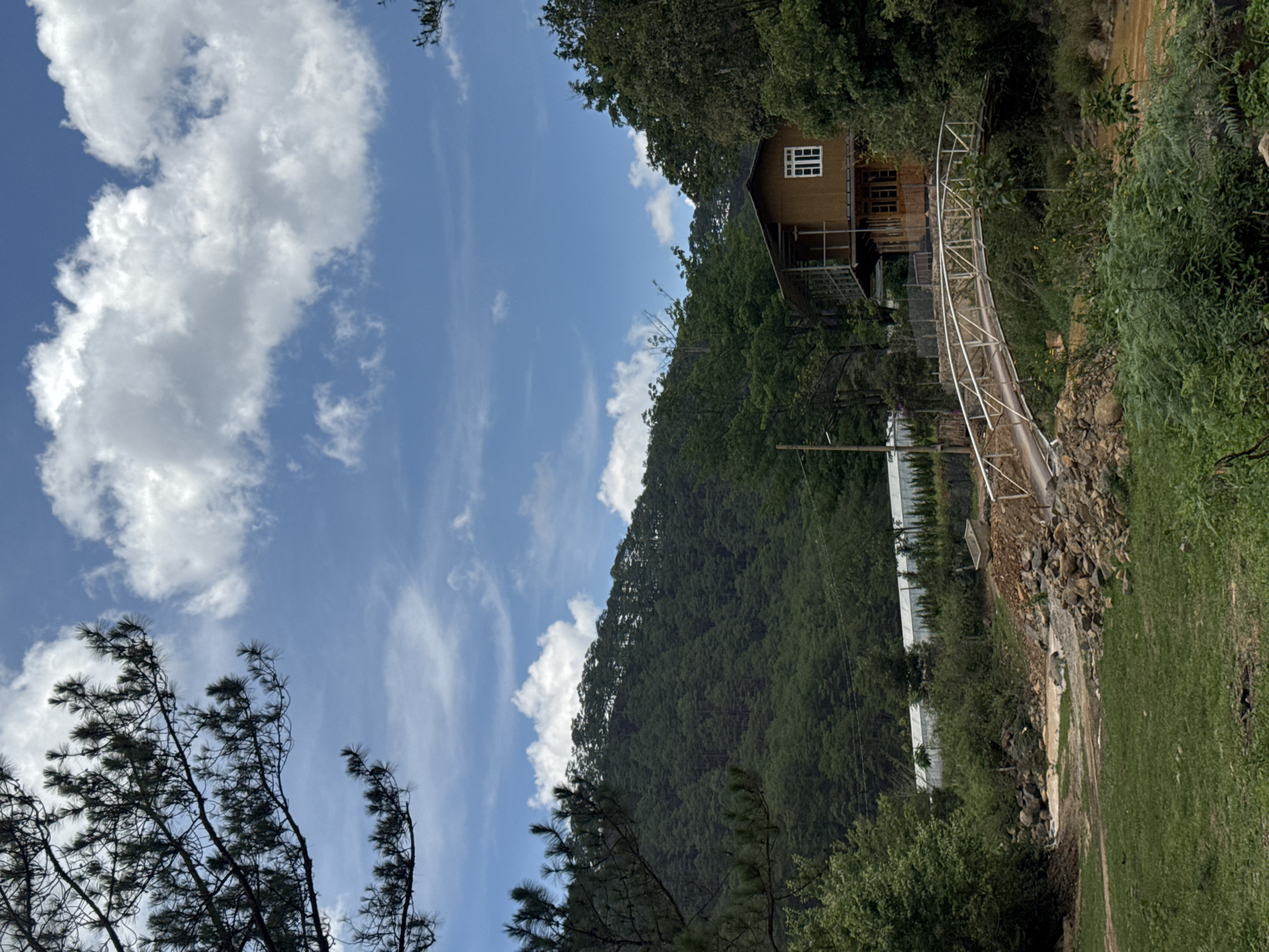 The scene is a rural area near Păng Tiêng 1, Vietnam. A wooden house with a white-framed window sits on a hillside, partially obscured by trees. A metal bridge leads from the house down the slope. Below the bridge is a rocky area with some construction. Beyond this, there are greenhouse-like structures. The sky is blue with scattered white clouds. In the foreground, there is grassy ground to the right and trees to the left.