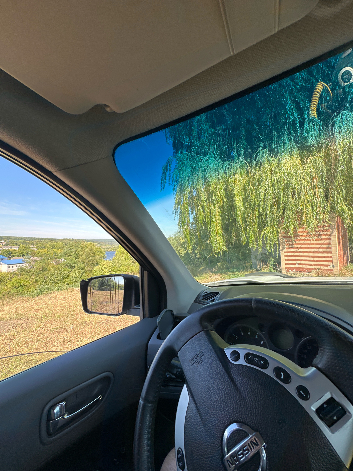 The image, taken from inside a car in Makiv, Ukraine, shows a view through the windshield and side window. The driver's side door panel and part of the steering wheel are visible in the foreground. Outside, a rural landscape unfolds with rolling hills covered in dry grass, scattered trees, and a distant body of water. A small settlement with blue-roofed buildings is visible on the left side. A wing mirror is attached to the driver's side door frame. The windshield is partially obscured by the branches of a weeping willow tree and a small, striped structure resembling a shed or outbuilding. A centipede-like insect is visible on the upper right portion of the windshield. The sky is clear and blue.