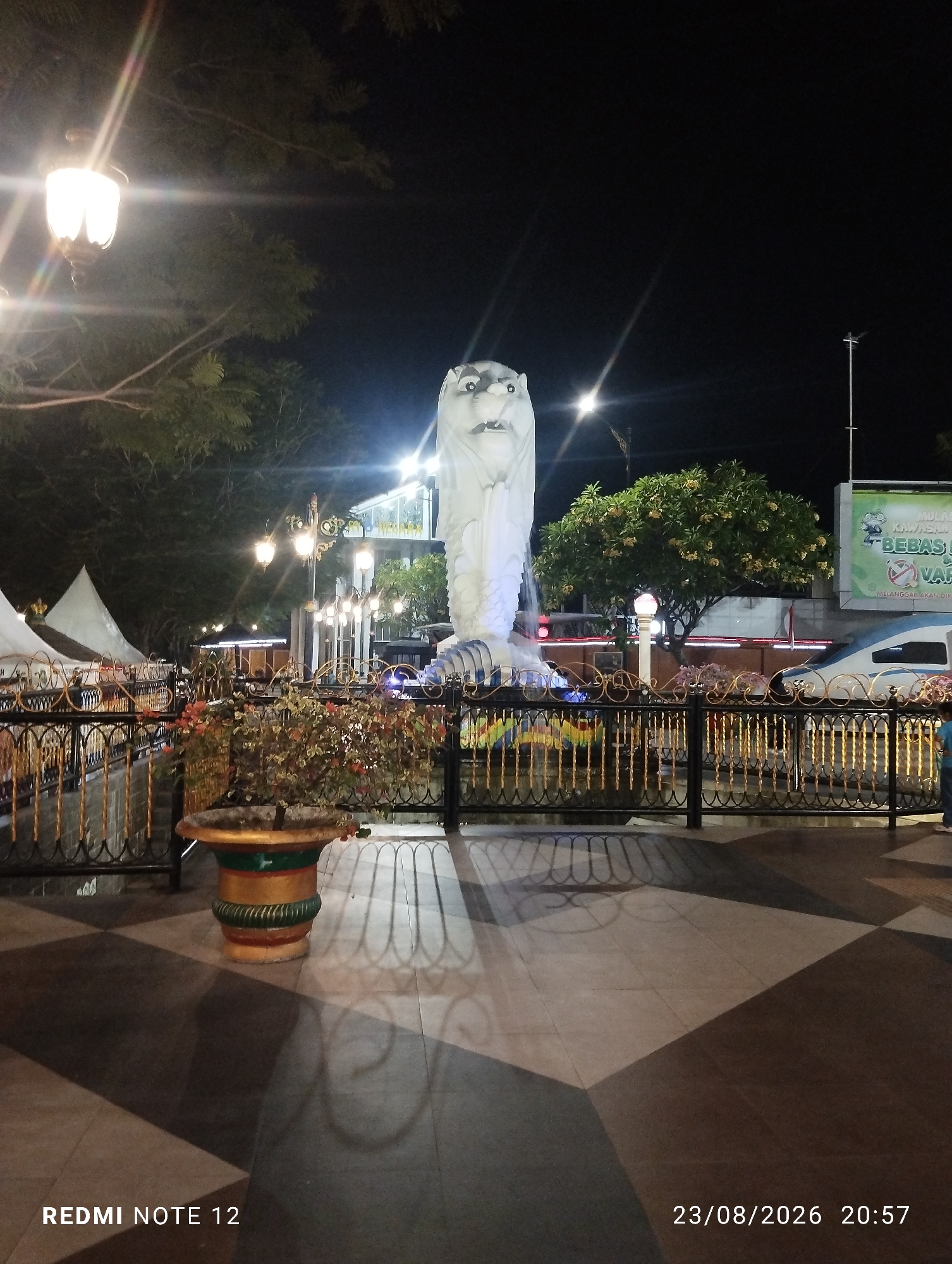 The image shows the Merlion statue in the city of Madiun, Indonesia, at night. The statue is a white, mythical creature with the head of a lion and the body of a fish, illuminated by various lights. It is surrounded by a decorative fence with golden accents. In the foreground, there is a paved area with a checkered pattern, and a planter containing plants. Several streetlights are visible, casting a warm glow. In the background, there are trees, tents, a building, and a vehicle. A banner with text is also visible in the background. The timestamp "23/08/2026 20:57" and "REDMI NOTE 12" are superimposed on the image.