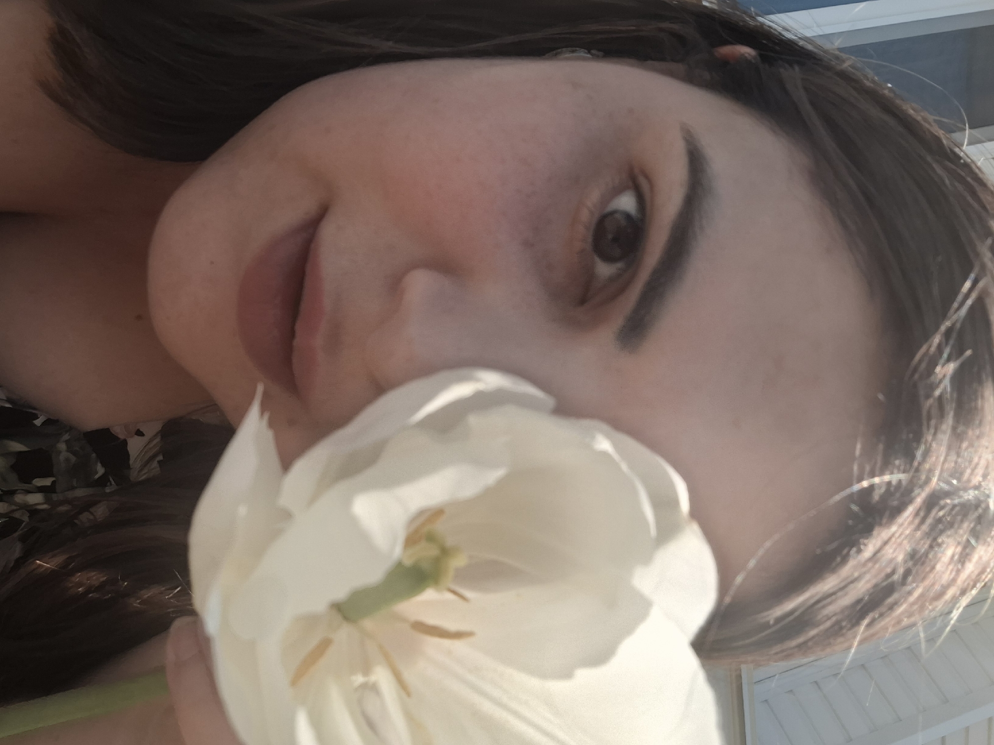 A close-up selfie shows a woman holding a white tulip flower near her face. Her hair is dark brown, and her skin is fair. The location is Амзя, Russia, inferred from the user request. The background shows partial view of a bright day, possibly outdoors. The interaction is the woman posing with the flower.