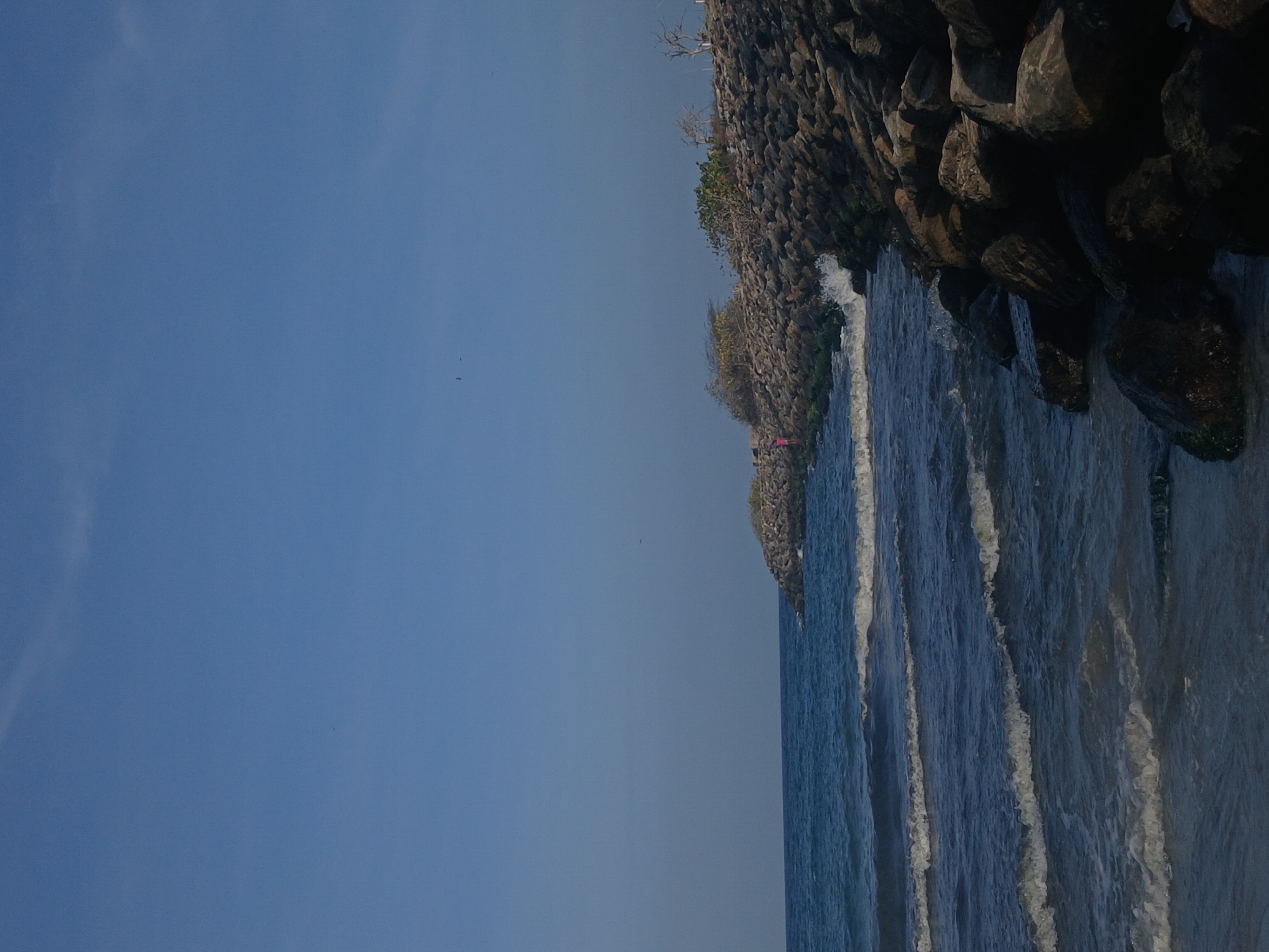The image shows a shoreline with a rocky cliff on the right and the ocean meeting the rocks. There's vegetation on the cliff. The sky is visible on the left, suggesting it's daytime. The location is Porlamar, Venezuela. I can see waves crashing against the rocky shore. There's no presence of people, interactions and actions in the image.