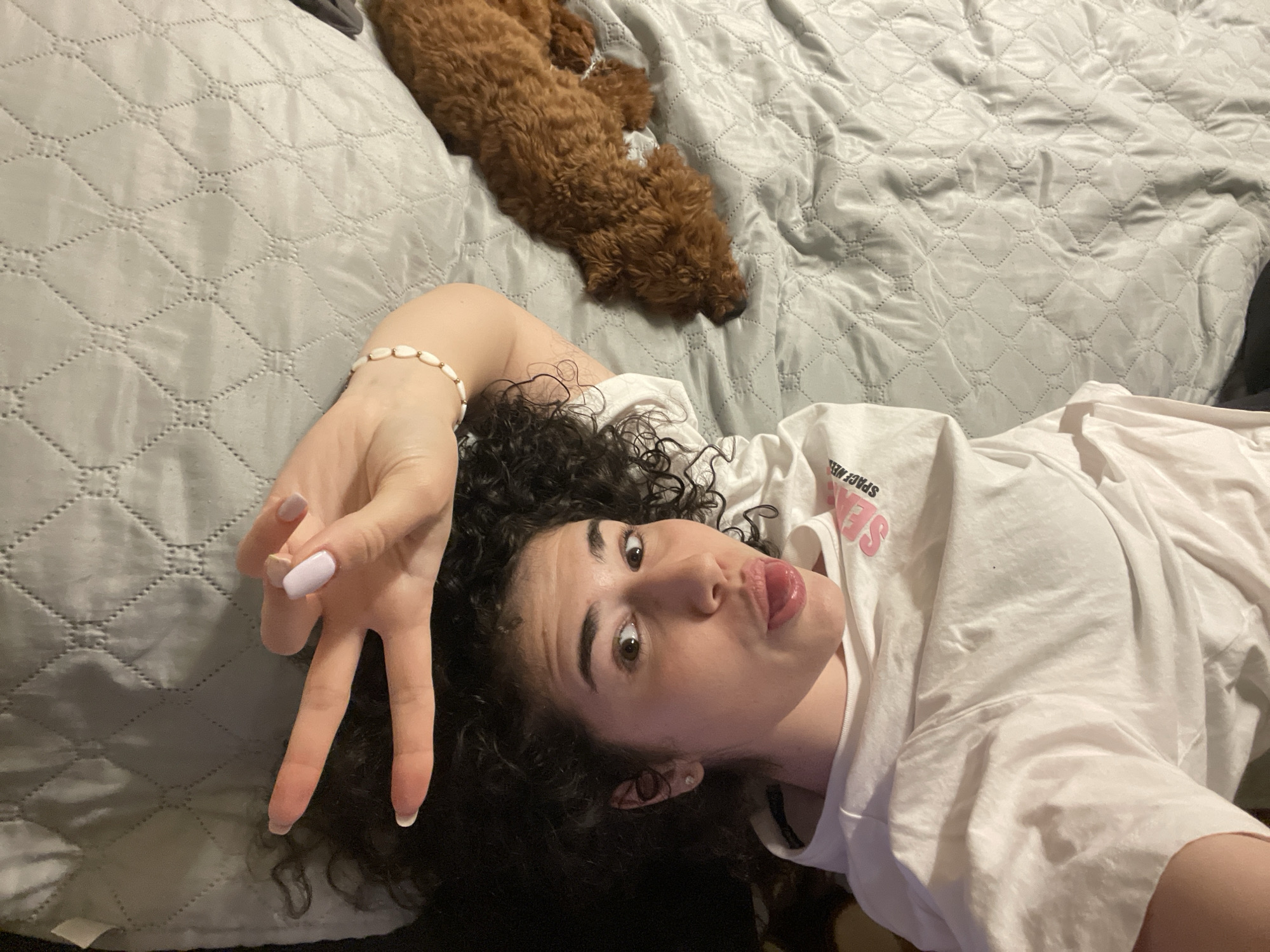 A young woman lies on a gray quilted bed, making a peace sign with her left hand. She has curly dark hair and is wearing a white t-shirt. Her tongue is slightly protruding. Behind her, a brown poodle is sleeping. The location is not discernible from the image.