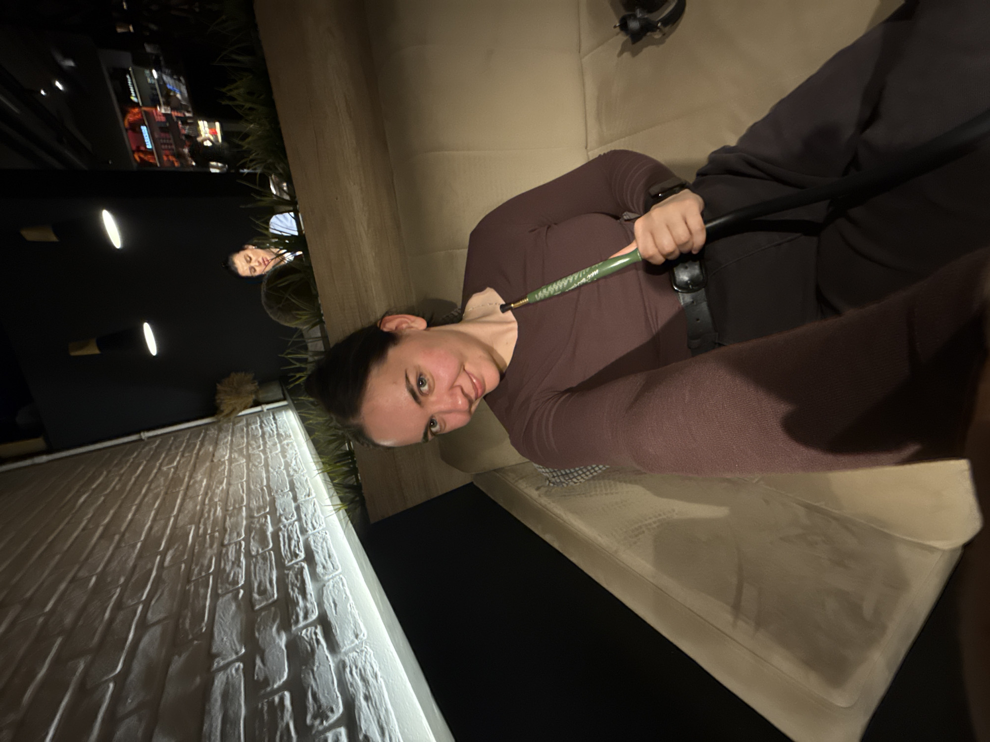 A person is holding a green and black object, possibly a hookah pipe, in an indoor setting. Another person is visible in the background. The location is Chaikovsky, Russia, as suggested by context, but there are no elements within the image itself that independently confirm this location. The indoor setting appears to be a casual or lounge area, possibly with outdoor access, suggested by a brick wall and night-time lighting visible in the background.