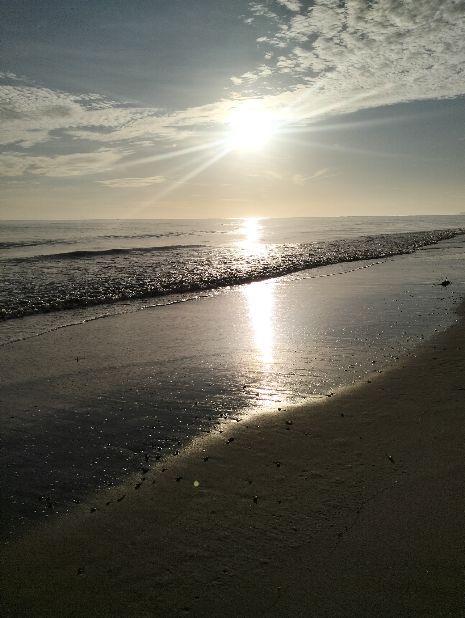 The image depicts a coastal scene at Sagoe, Indonesia. The sun is positioned high in the sky, obscured by thin clouds, creating a bright glare and long reflection on the water and wet sand. Waves are visible, approaching the shore. The beach stretches into the foreground, transitioning from wet, reflective sand to dry sand. There are few objects visible on the beach.