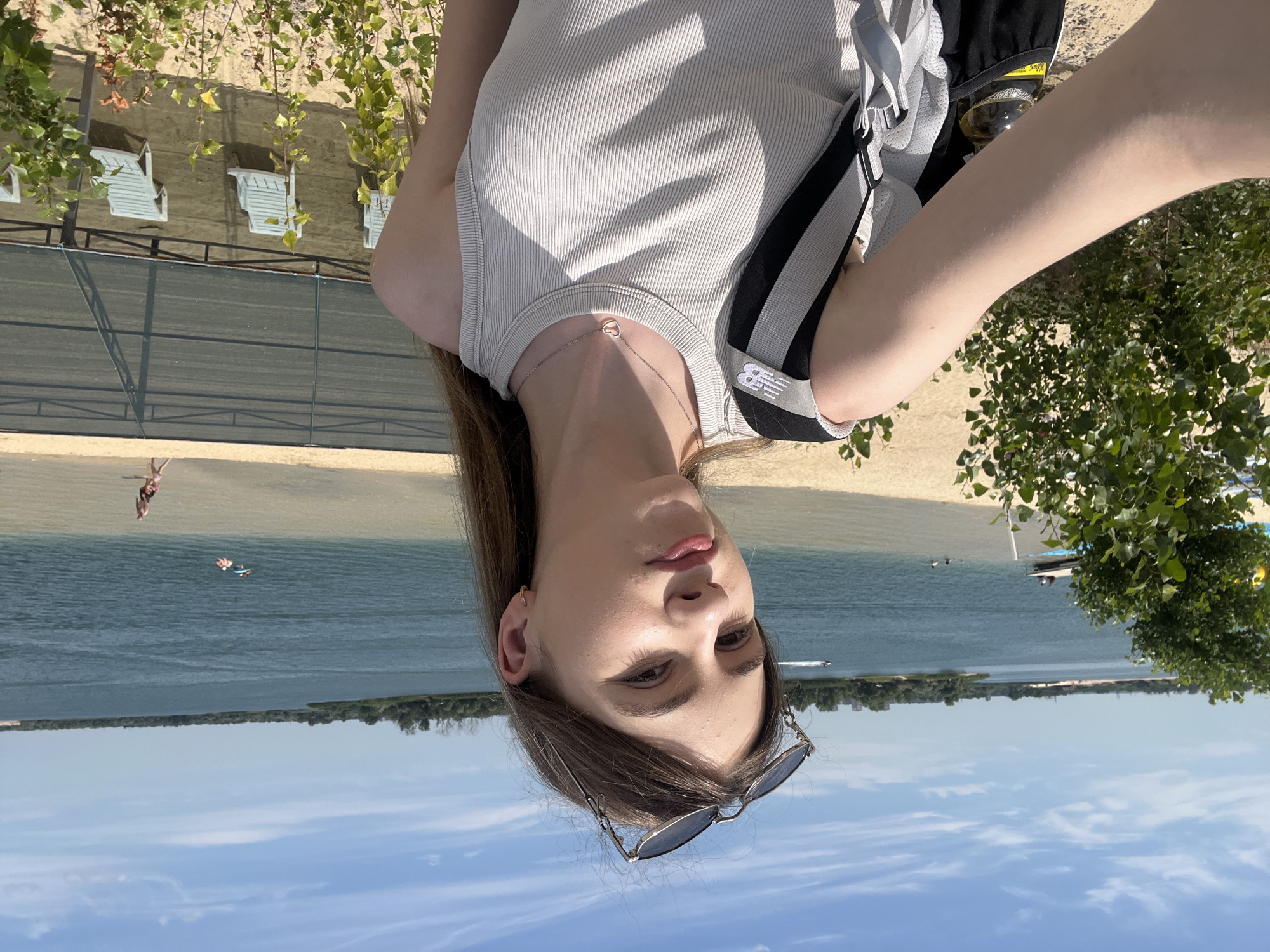 The media file shows a young woman taking a selfie outdoors. She is wearing a grey ribbed tank top and sunglasses perched on her head. The background reveals a beach with lounge chairs and trees. Beyond the beach, a body of water with a few small boats and a distant tree line is visible. The sky is blue with scattered clouds. The image appears to be taken during the daytime, likely on a sunny day. The woman's expression is neutral. There is no visible text in the image. The location context is Yelyzavetivka, Ukraine.