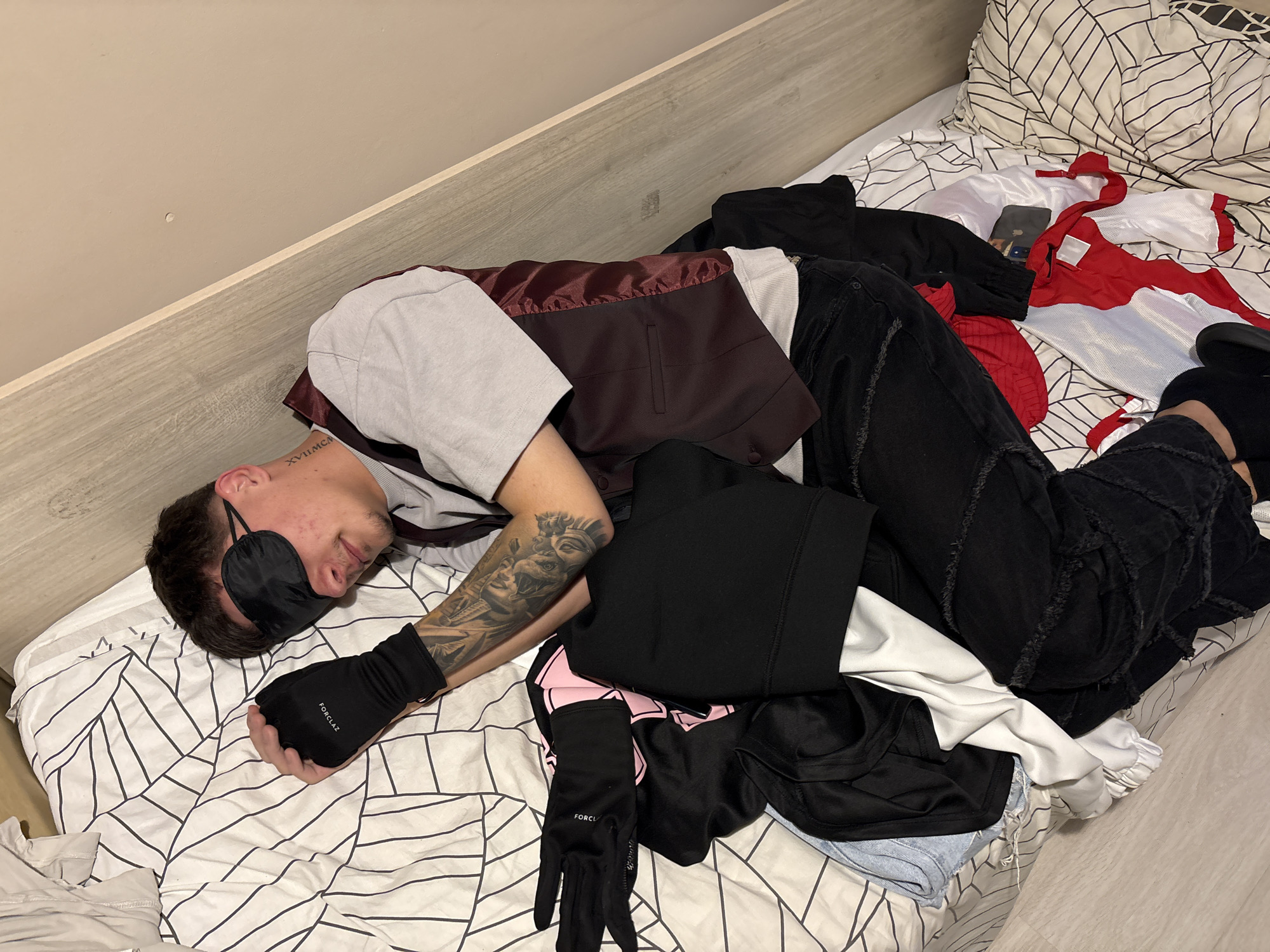 The image captures a young man asleep or resting on a bed within an indoor bedroom setting, located in Plovdiv, Bulgaria. The man is positioned on his right side, facing slightly towards the camera, with his head on a white duvet cover adorned with a black abstract geometric pattern. He is wearing a black sleep mask over his eyes, a light gray short-sleeved t-shirt, and a maroon vest. His lower body is clad in dark (likely black) ripped jeans. A black fingerless glove, with the brand "FORCLAZ" printed on it, covers his left hand. A detailed tattoo of what appears to be a classical sculpture or figure is prominent on his left forearm. Another tattoo, "XVIIIMCM", is visible on the right side of his neck, just below his ear.
The bed is situated next to a light-colored wooden headboard or wall paneling, with a plain light beige wall visible above. The bed area is cluttered with various articles of clothing, including a red and white jacket-like garment, a black hoodie or sweatshirt, and other dark and light fabric items. A smartphone, encased in a dark cover, is partially visible amongst the clothes near the man's feet. The indoor lighting is neutral, and the presence of a sleep mask suggests either daytime sleep or an effort to block out light during rest. The man's posture indicates a state of relaxation or sleep.