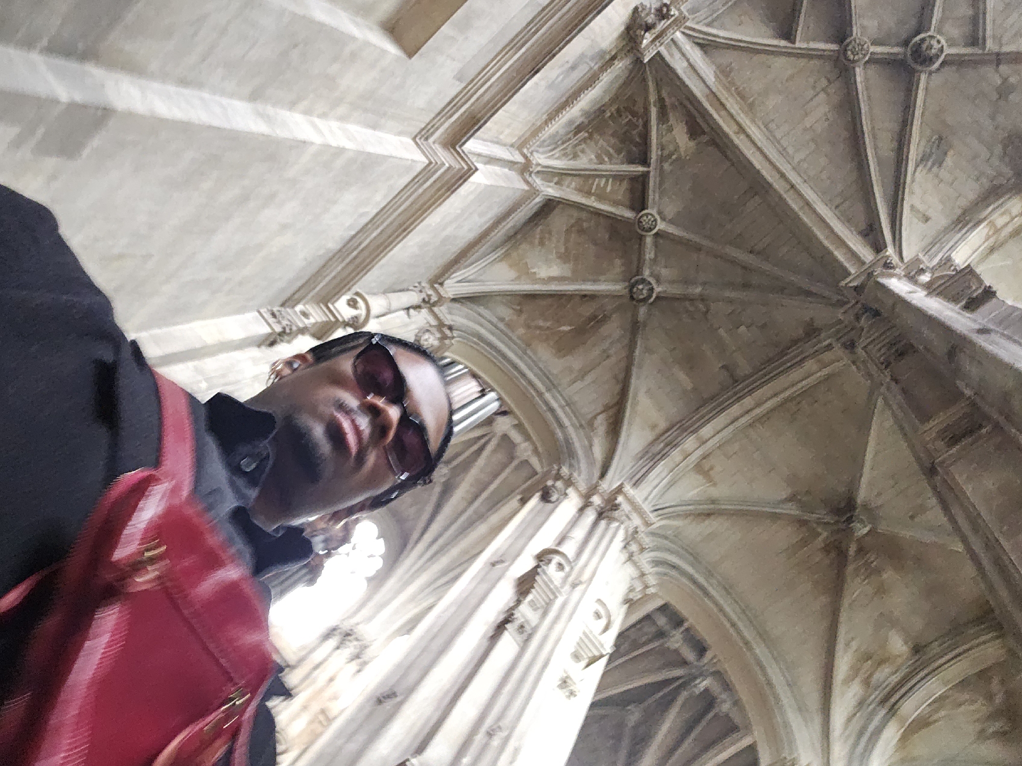 Here is a detailed description of the image:
**Scene:**
The image captures a low-angle perspective of a man inside a Gothic architectural space. The focus is shared between the man and the intricate ceiling design.
**People:**
A Black man is visible, primarily from the chest up. He is wearing a black shirt and sunglasses. A red backpack is slung over his shoulder.
**Environment:**
The setting is a cathedral or similar Gothic building, characterized by vaulted ceilings with ribbed arches. Stone pillars and archways frame the background. The building appears old with aged stonework.
**Action/Interaction:**
The man appears to be taking a photograph, as the camera angle suggests a selfie or photo taken from a low perspective. He is looking directly at the camera.
**Location:**
Given the Gothic architecture, it is very likely the man is inside Sainte-Chapelle in Paris, France.