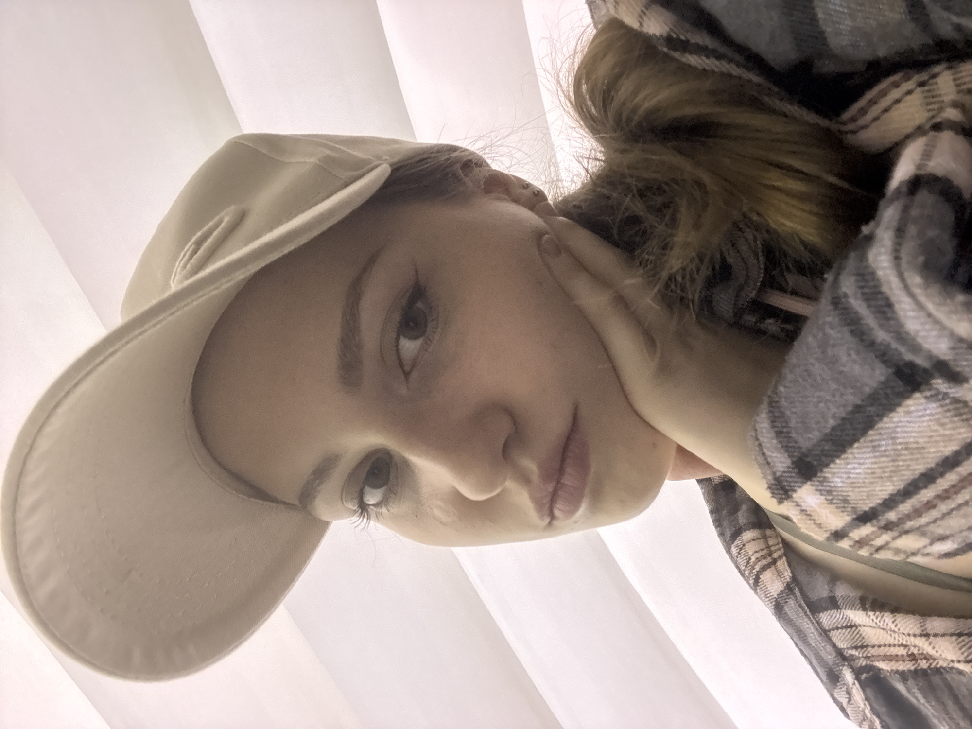 A young woman with a beige baseball cap tilted forward is shown in a close-up selfie. She is resting her chin on her right hand, with her fingers fanned across her cheek. Her left eye is visible, with defined eyeliner. She is wearing a plaid shirt with shades of grey, white, and pink. Her hair, which is a light brown color, is visible pulled back. The background consists of white, corrugated blinds. The image does not contain any identifiable elements that would indicate a specific location in Saint Petersburg, Russia.