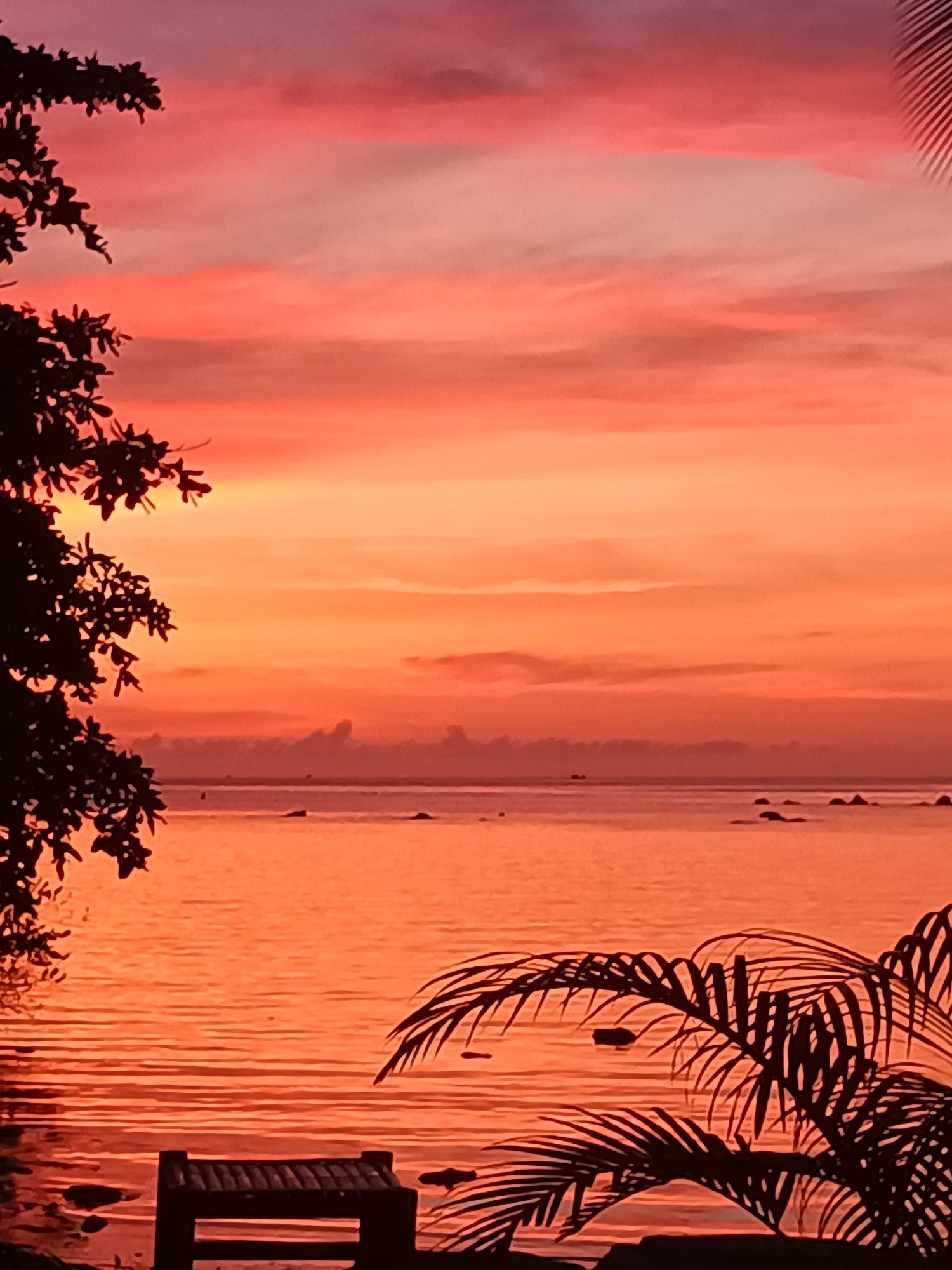 A scenic image depicts a tranquil coastal scene at Baan Sri Thanu, Thailand. The sky exhibits a vivid gradient of orange and pink hues, typical of a sunset or sunrise. The ocean reflects the warm colors of the sky, creating a mirrored effect. Silhouetted trees and palm fronds frame the edges of the image, adding depth and contrasting the bright sky. In the foreground, there's a small, dark structure, possibly a wooden dock or bench, positioned near the water's edge. The overall atmosphere is serene and tropical.