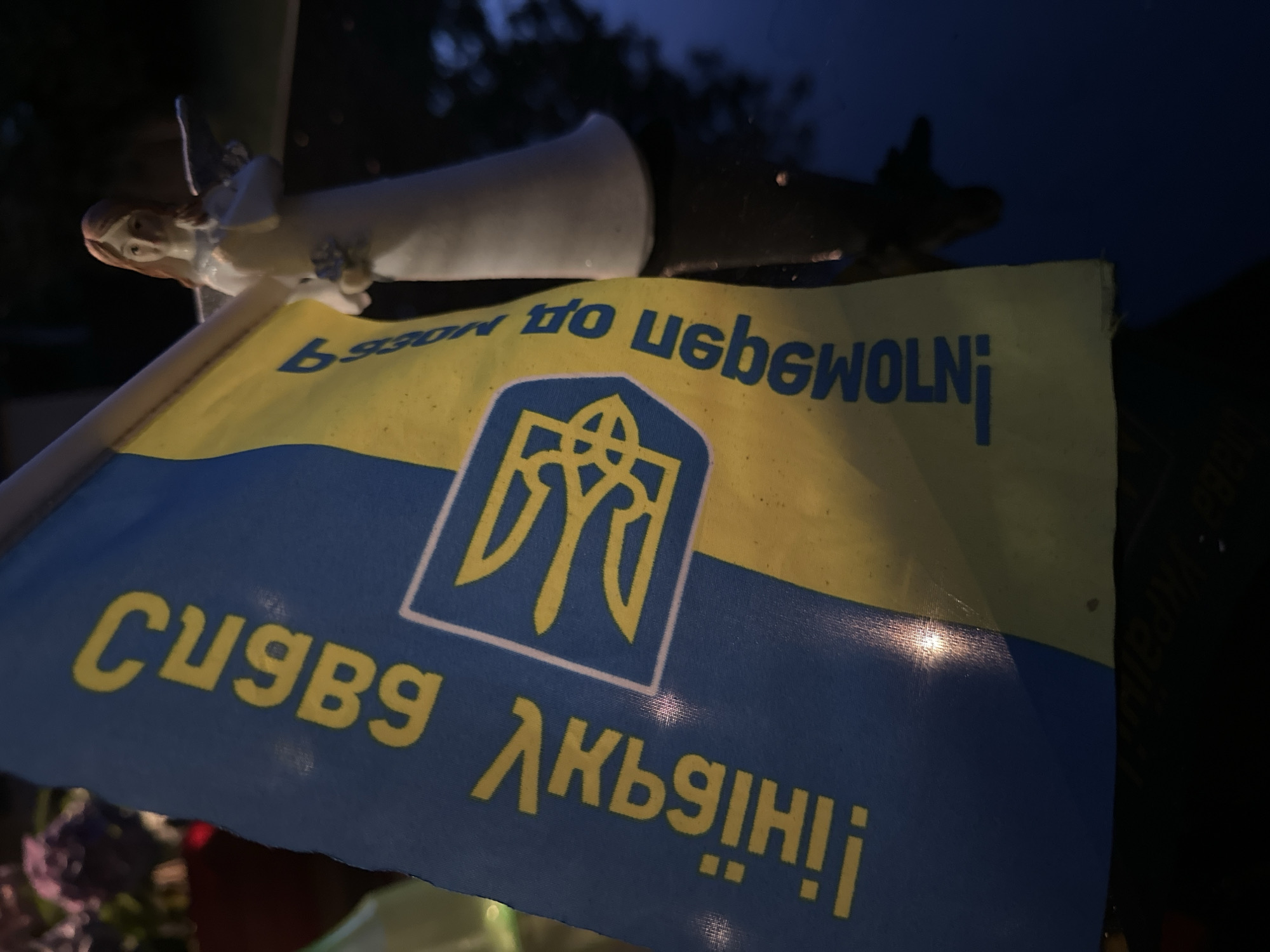 An image shows a Ukrainian flag with text in yellow. The flag displays the Tryzub, the coat of arms of Ukraine, a golden trident on a blue shield. To the right of the flag is a white angel figurine with wings and blonde hair, holding a small bouquet of flowers in its hands. The figurine is positioned in front of a dark, reflective surface, possibly a car hood or table. In the background, blurred purple flowers are visible, suggesting an outdoor or garden setting. The overall lighting is dim, creating a moody atmosphere. The photograph was taken in Krefeld, Germany.
