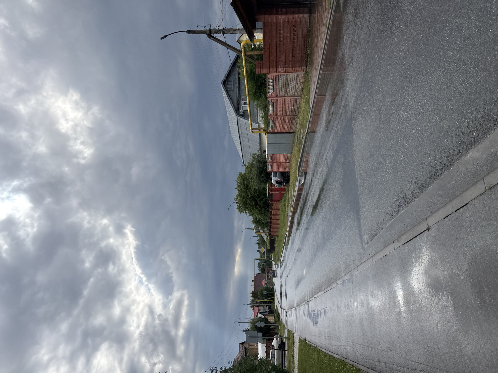 This image depicts a wet street scene in Kazan, Russia, likely after a rain shower. The overcast sky, with dramatic, textured clouds, suggests the weather is either still inclement or has recently cleared. A bright spot in the clouds hints at the possibility of the sun breaking through, though the overall mood is subdued and grey.
The scene is a residential street with houses and fences lining one side. The houses vary in style, with some featuring brick and others metal siding. A prominent yellow gas pipe runs along the side of one building and extends onto the street. Streetlights are visible, indicating it is likely daytime, but the dim light suggests it could be early morning or late afternoon.
The wet asphalt road reflects the grey sky and surrounding buildings, creating a glossy, almost mirror-like surface. Puddles are visible, further emphasizing the recent rain. A curb separates the road from a strip of green grass, beyond which lie the properties. A few cars are parked along the street or in driveways, with one white car prominently visible. The overall impression is one of a quiet, ordinary day in a residential area, marked by the aftermath of rain. No people are clearly visible in the image. There is no discernible text.