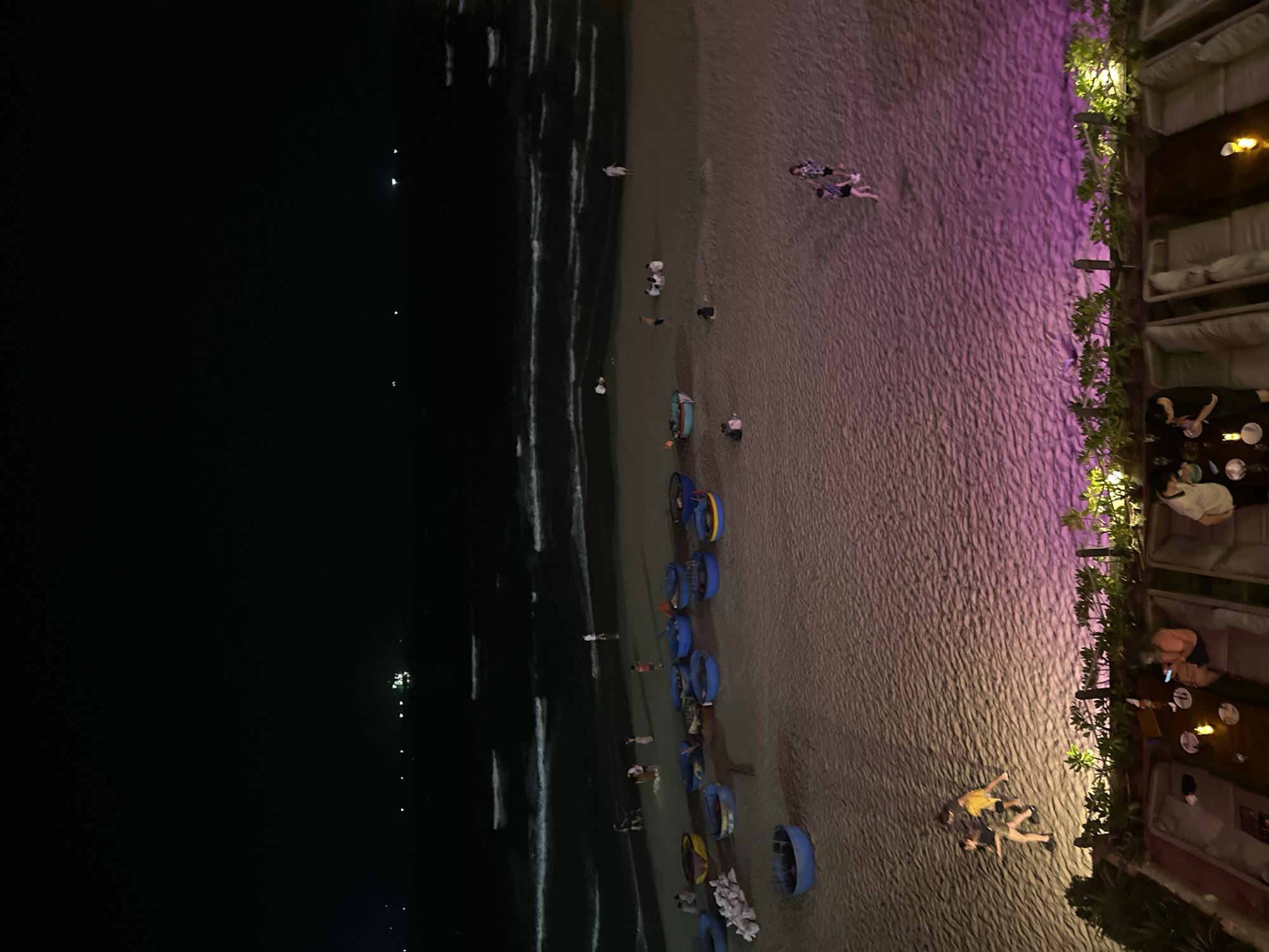 The image depicts a nighttime scene at My Khe Beach in Đà Nẵng, Vietnam. On the left, the dark expanse of the sea is visible, with faint light reflecting off the waves. A sandy beach stretches across the middle, where several people are strolling or lying down. A row of coracles is parked at the edge of the water. On the right side of the image, there is a row of beachside bars or restaurants with lighting, where people are sitting, eating, and talking at tables.