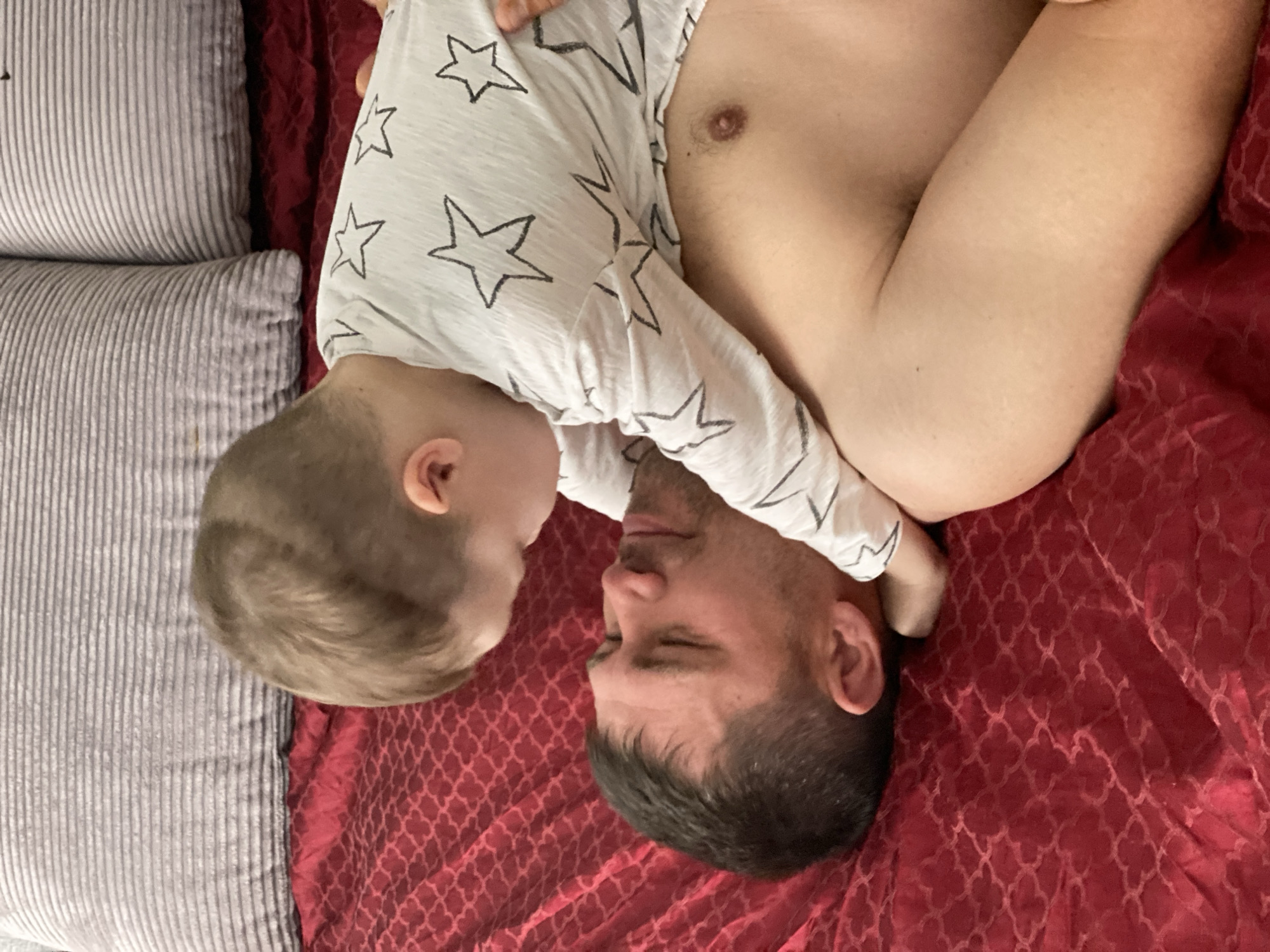 A man and a young child are lying on a bed with a red patterned bedspread. The man is lying on his back with his eyes closed, and the child is lying on top of him, face-to-face. The child is wearing a long-sleeved shirt with stars printed on it. There are grey striped pillows next to them. The location of Sochi, Russia is not discernible in the image.