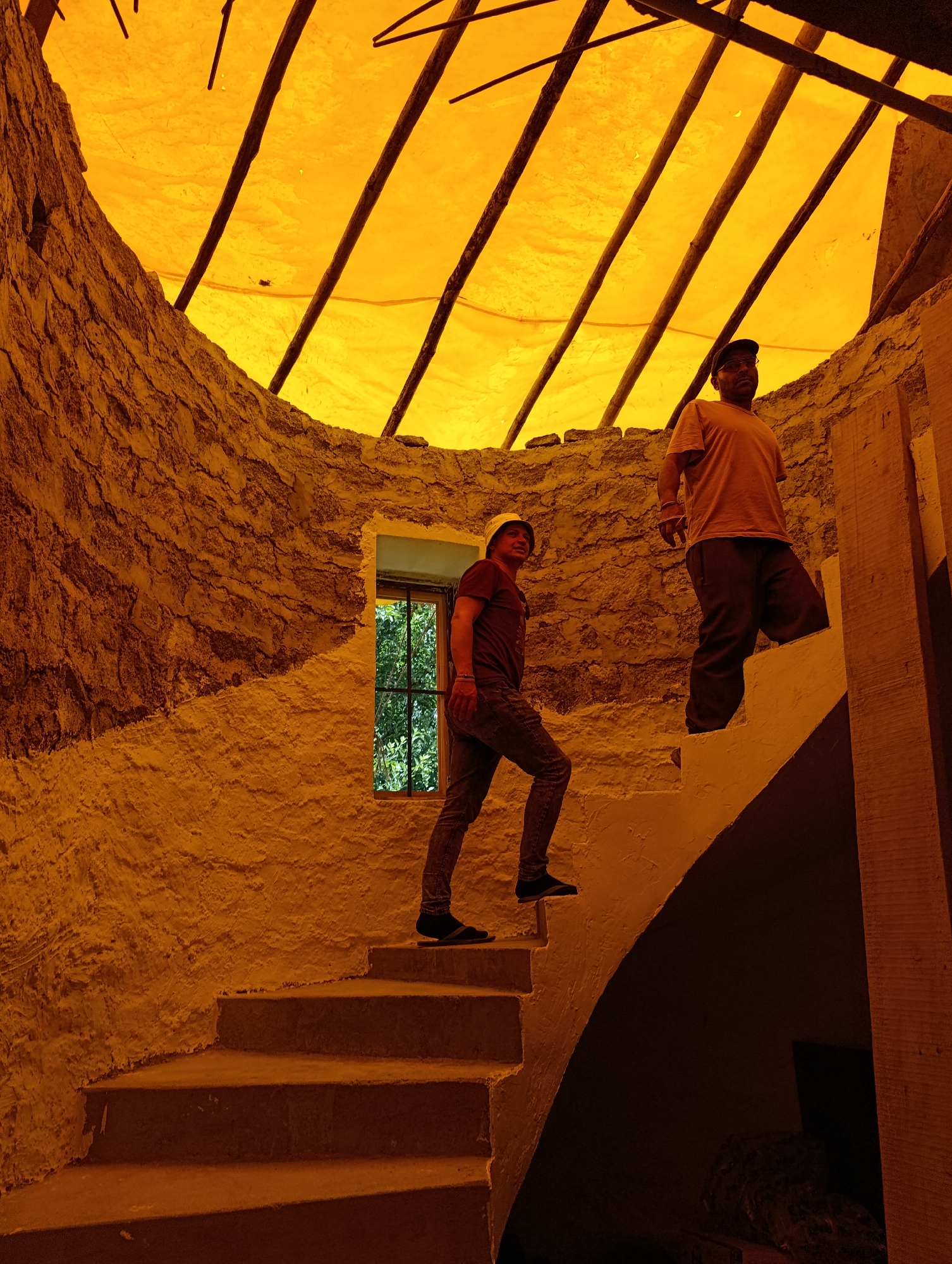 Two men are ascending a circular stone staircase inside a structure in Shanag, India. The walls are rough-hewn stone, partially covered with white plaster. A narrow window with a grid is visible on the left wall, looking out onto green foliage. The ceiling is an orange tarpaulin stretched over wooden beams. The man on the lower staircase wears a dark t-shirt and jeans, and a light-colored hat. The man on the upper staircase wears a light orange t-shirt, dark trousers, and a dark cap. The lighting is warm and orangish, likely from the tarpaulin filtering the sunlight. The stairs are made of concrete or a similar material.