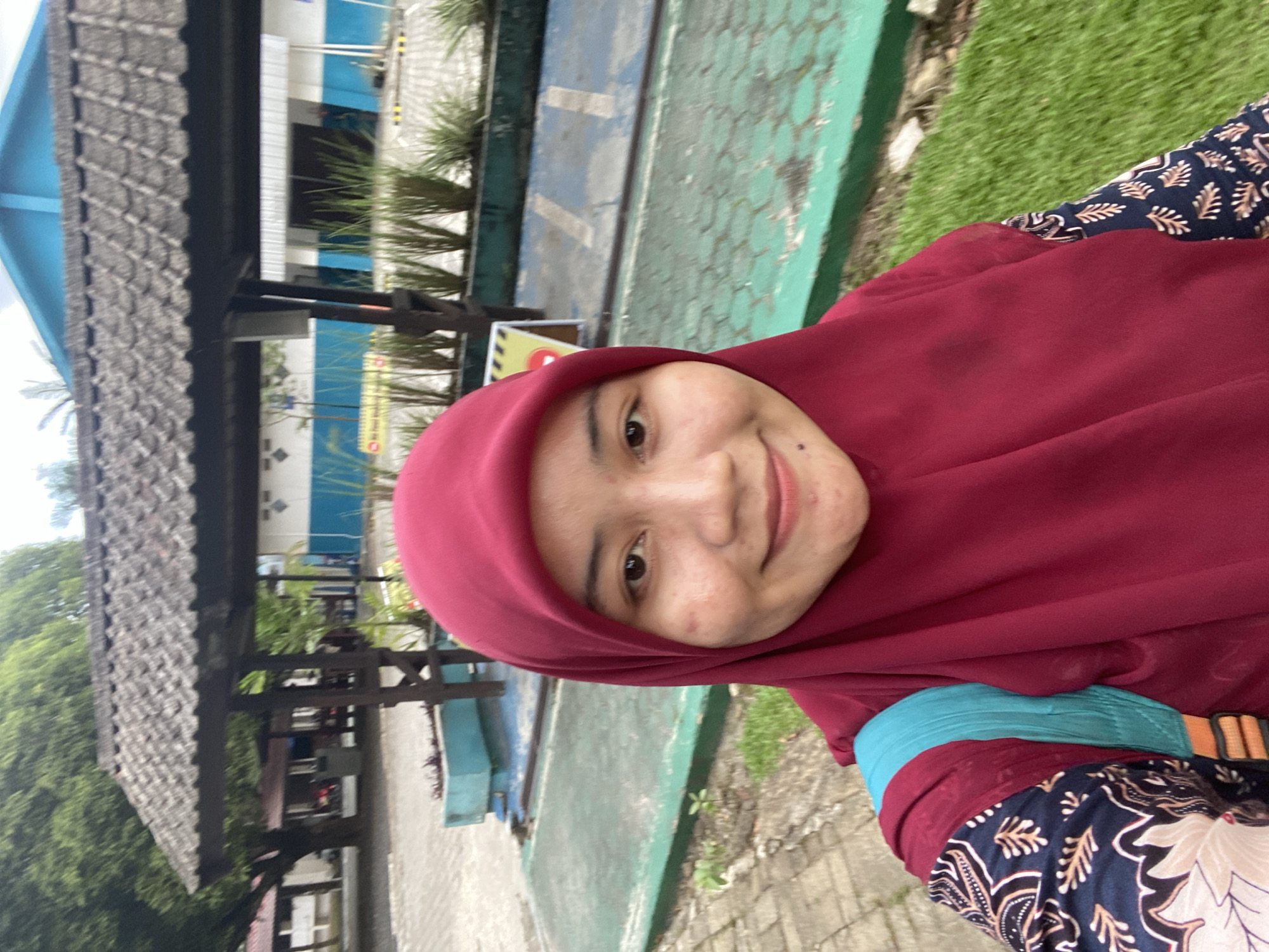 A young Indonesian woman wearing a red hijab and a patterned shirt is taking a selfie outdoors in a paved area with green grassy patches and some trees. Behind her is a low roofed structure with a tiled roof. Further in the background, a blue and white building is visible with some tall green plants in front of it. There is a yellow sign visible in the background. This scene is located in Banjarbaru, Indonesia.