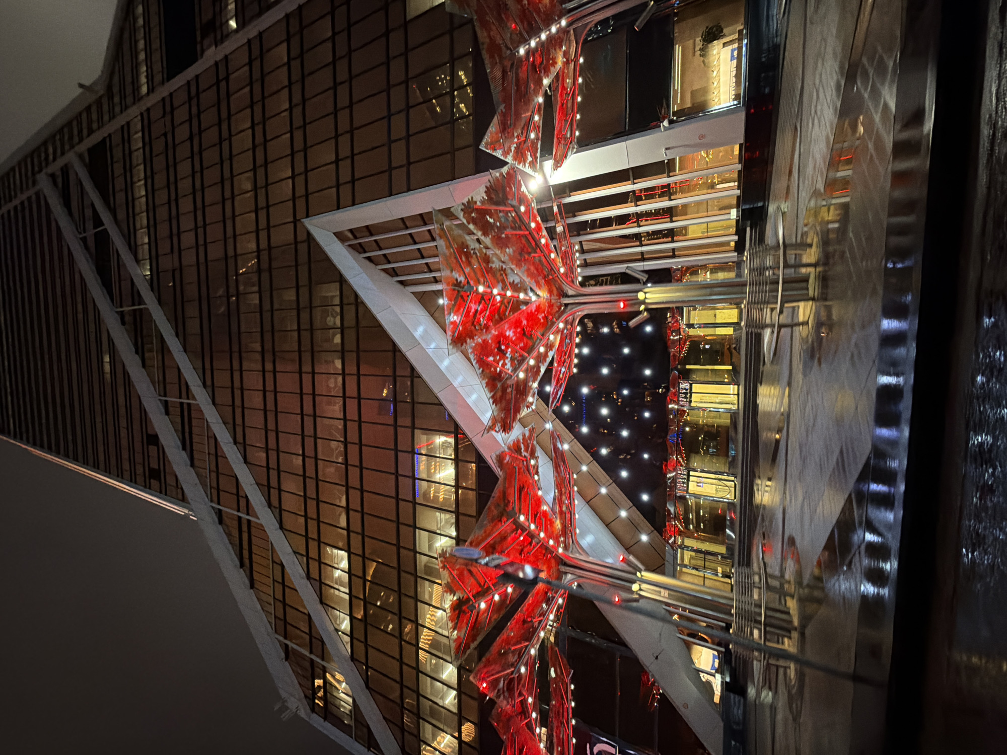 A night-time, low-angle photograph captures the architecture of a building decorated with red, geometrical patterns. The structure features a glass facade with visible gridlines. The red decorations are positioned within a triangulated framework, possibly part of a festive installation. The building's reflection is slightly blurred, suggesting the presence of a reflective surface. No people are visible. The location is likely a commercial or public area in an urban setting. The image does not definitively indicate it is located in the city of Khimki, Russia.