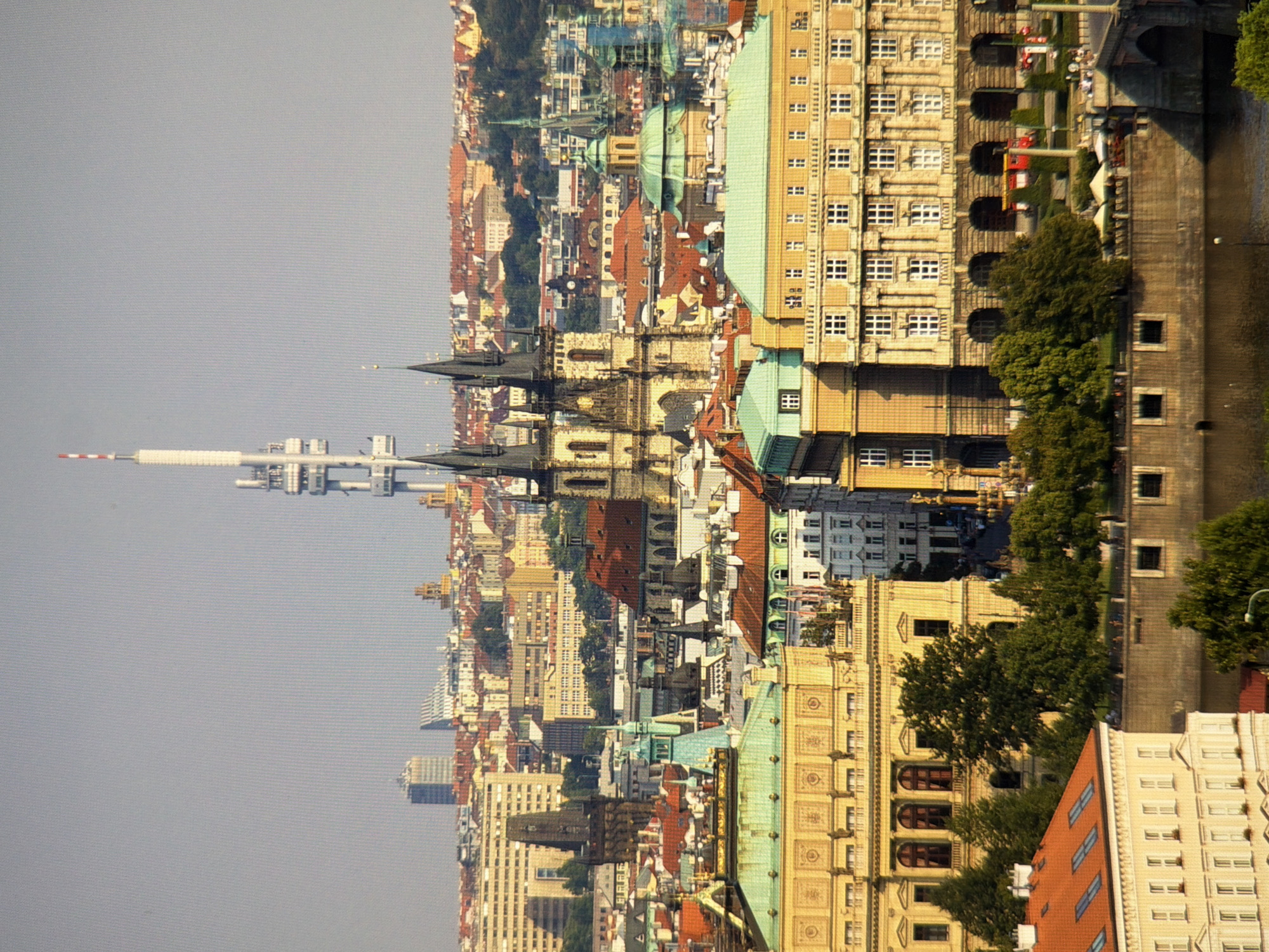 The image is an aerial view of a city skyline featuring a mix of architectural styles, including historical buildings with red-tiled roofs and spires, as well as more modern structures. A prominent television tower with a distinctive antenna structure stands tall in the background. In the foreground, a large, light-colored building with arched windows and ornate detailing is visible, adjacent to a building with a bright orange-tiled roof. Trees and a body of water, likely a river, are also present. The sky is clear and pale. The scene appears to be a daytime shot with sunlight illuminating the cityscape. This is a view of Prague, Czech Republic, not Kyiv, Ukraine.