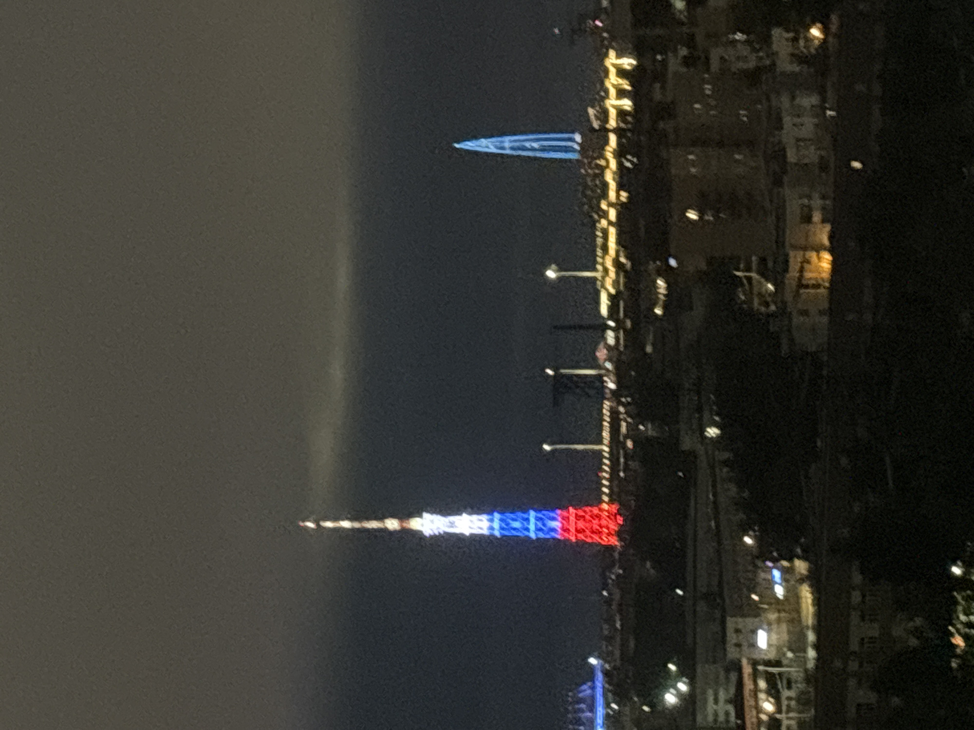 The image shows a nighttime view of a city. The foreground and background are dark, while the center features illuminated structures. One of the most prominent features is a tall tower lit in red, white, and blue. Another, less defined structure appears in the upper part of the image, also illuminated in blue. Other lights from buildings and street lamps can be seen scattered throughout the scene. The image is grainy. The location appears to be an urban area, likely at night.
