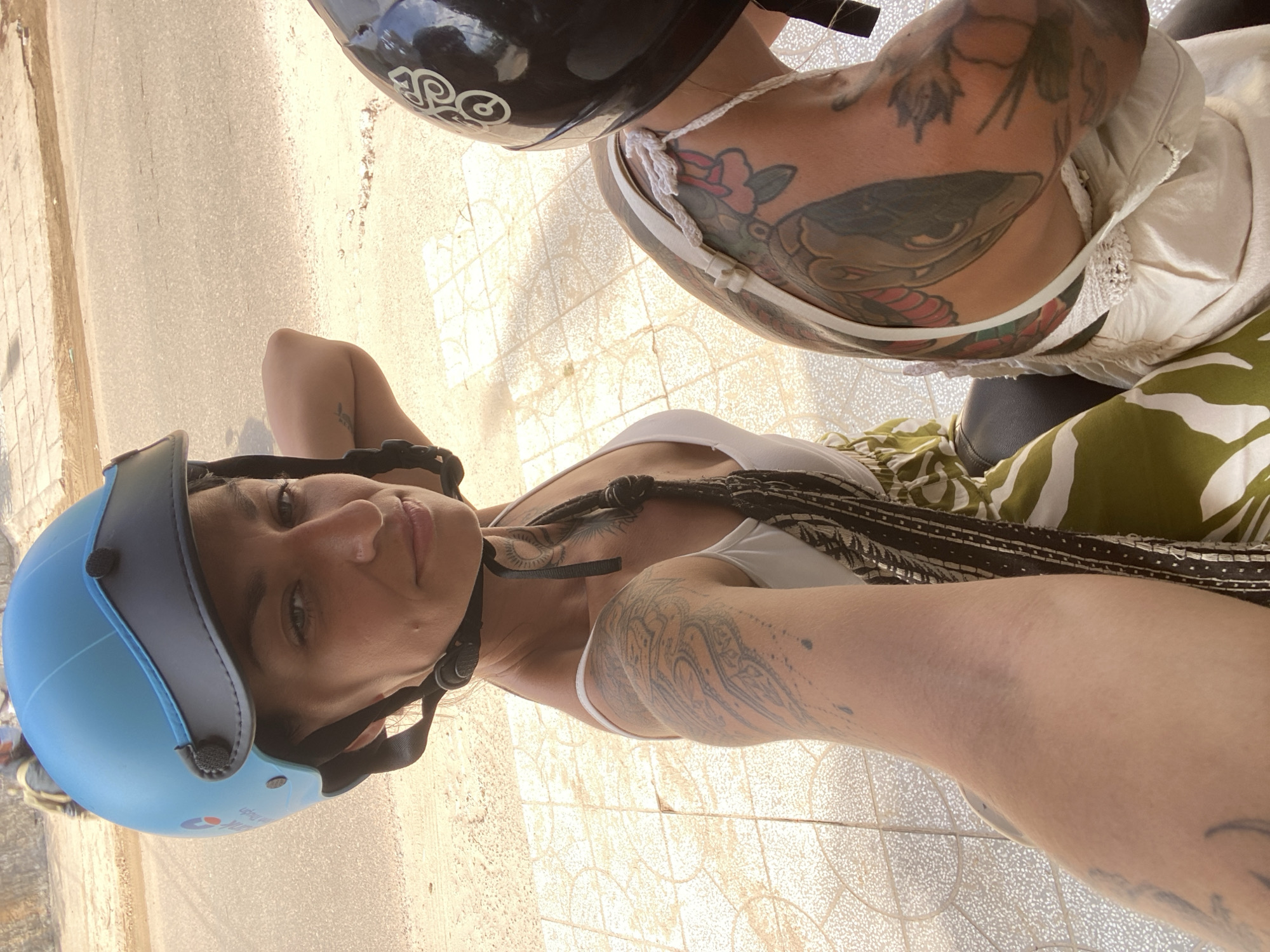 In Mũi Né, Vietnam, two women are riding on a motorbike. The woman in the foreground, wearing a blue helmet, is taking a selfie. She has tattoos on her arm and shoulder. She is wearing a white tank top and a patterned skirt. The woman in the background is wearing a black helmet and has tattoos on her back. The motorbike is on a paved road, with a sidewalk to the left. The sun is shining, casting shadows on the ground. The environment appears to be urban, with hints of sandy terrain in the background.