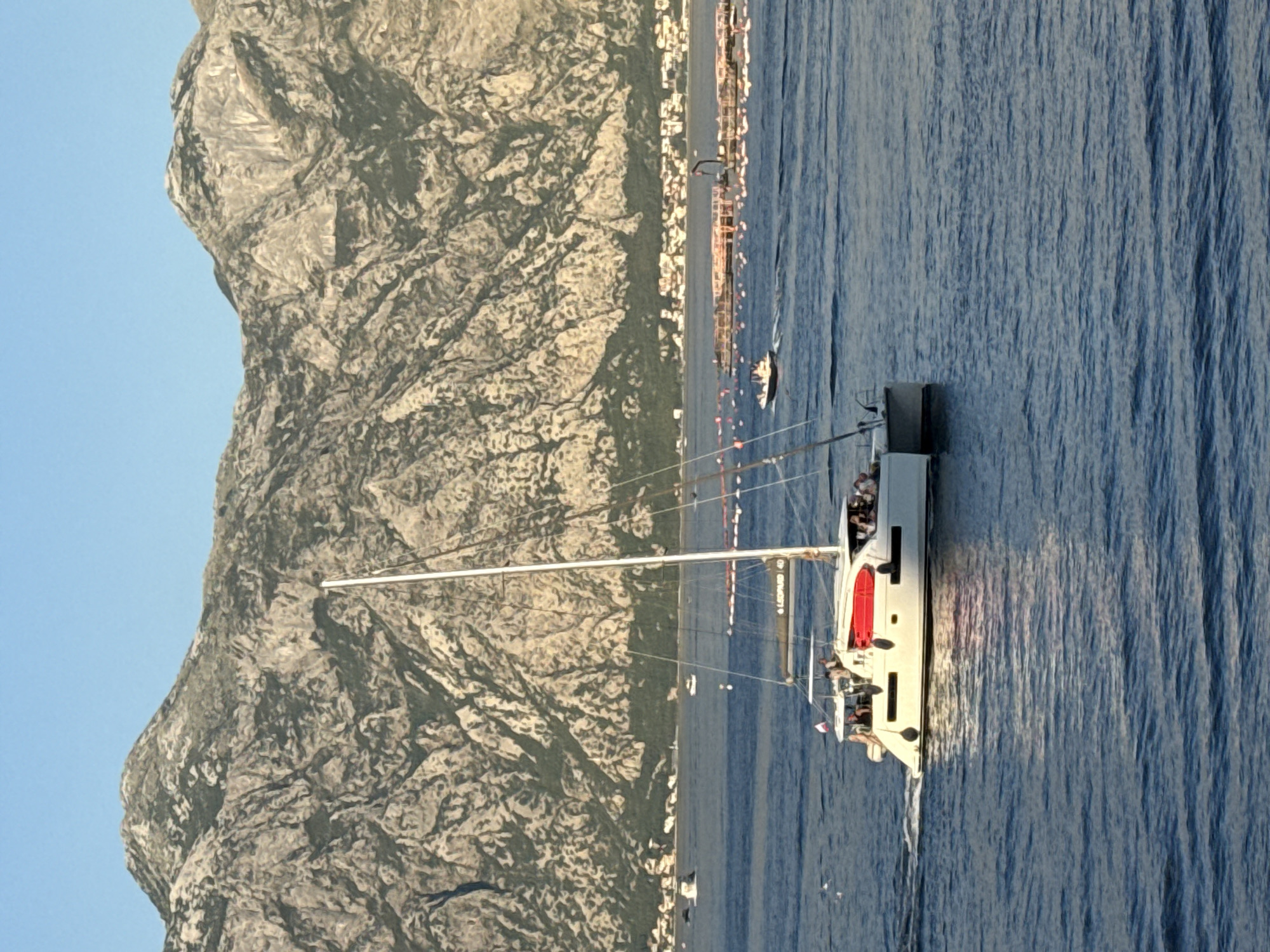 The image captures a catamaran sailing on a body of water with a large, mountainous terrain in the background. The catamaran, white with red accents, is populated by several individuals. In the middle ground, a smaller boat is visible, and further in the distance, a network of buoys and a crane suggest some sort of marine infrastructure or aquaculture. The sky is clear and blue. This scene is likely located in Đurići, Montenegro, given the characteristic mountainous coastline of the region.