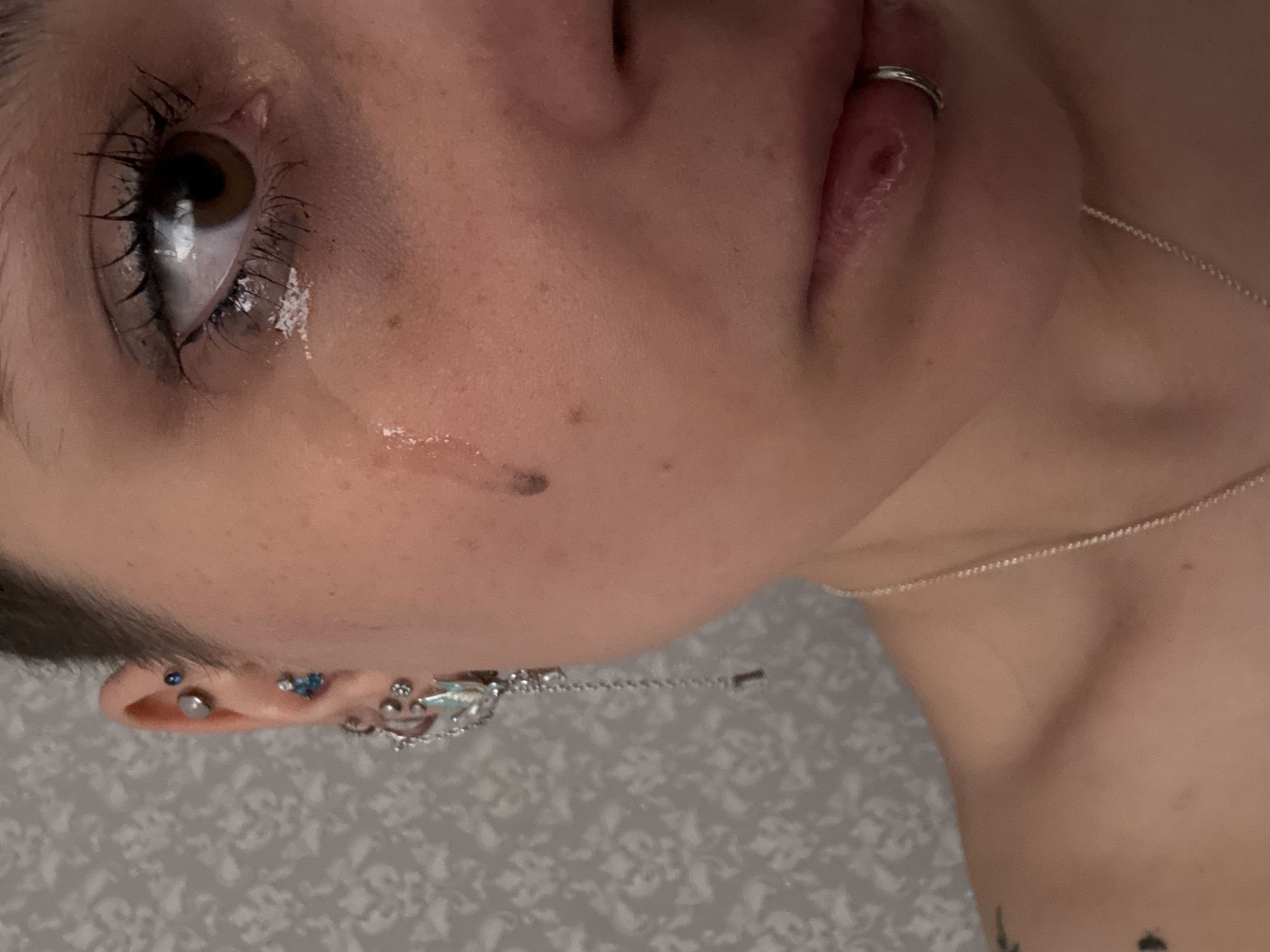 A close-up, slightly angled portrait shows a person with fair skin, dark eyelashes, and a brown eye with a distinct white highlight. A clear liquid, possibly a tear, is visible on the lower eyelid and a trail of it runs down the cheek. The person has a lip piercing with a silver ring. The ear has multiple piercings, including a row of small blue gems. A silver chain necklace is visible on the person's chest. The background is a patterned surface, possibly a wall or fabric. The image does not indicate the location of Saint Petersburg, Russia.